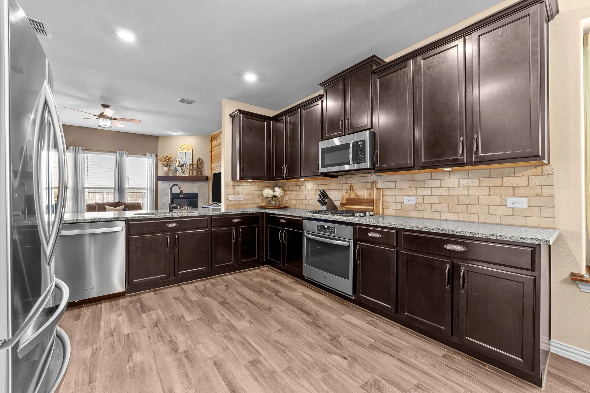 Modern kitchen featuring dark cabinetry with stainless steel hardware, stainless steel appliances including a built-in microwave and wall oven, subway tile backsplash, granite countertops, and wood-finish tile flooring throughout the open layout