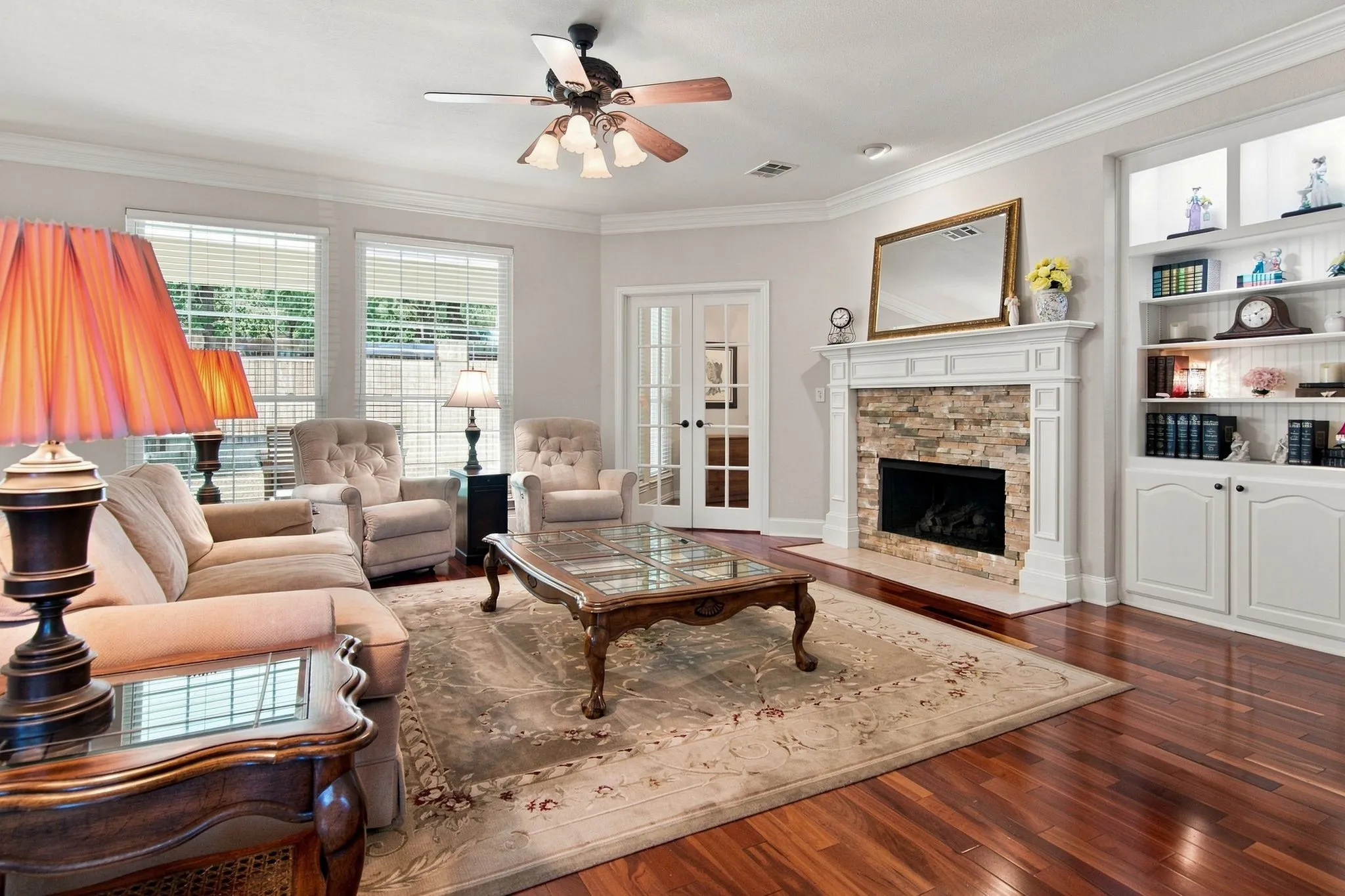 Elegant living room showcasing hardwood flooring, a wood-burning fireplace with decorative surround and mantel, built-in shelving with cabinet storage, a ceiling fan with integrated lighting, and expansive windows with natural light