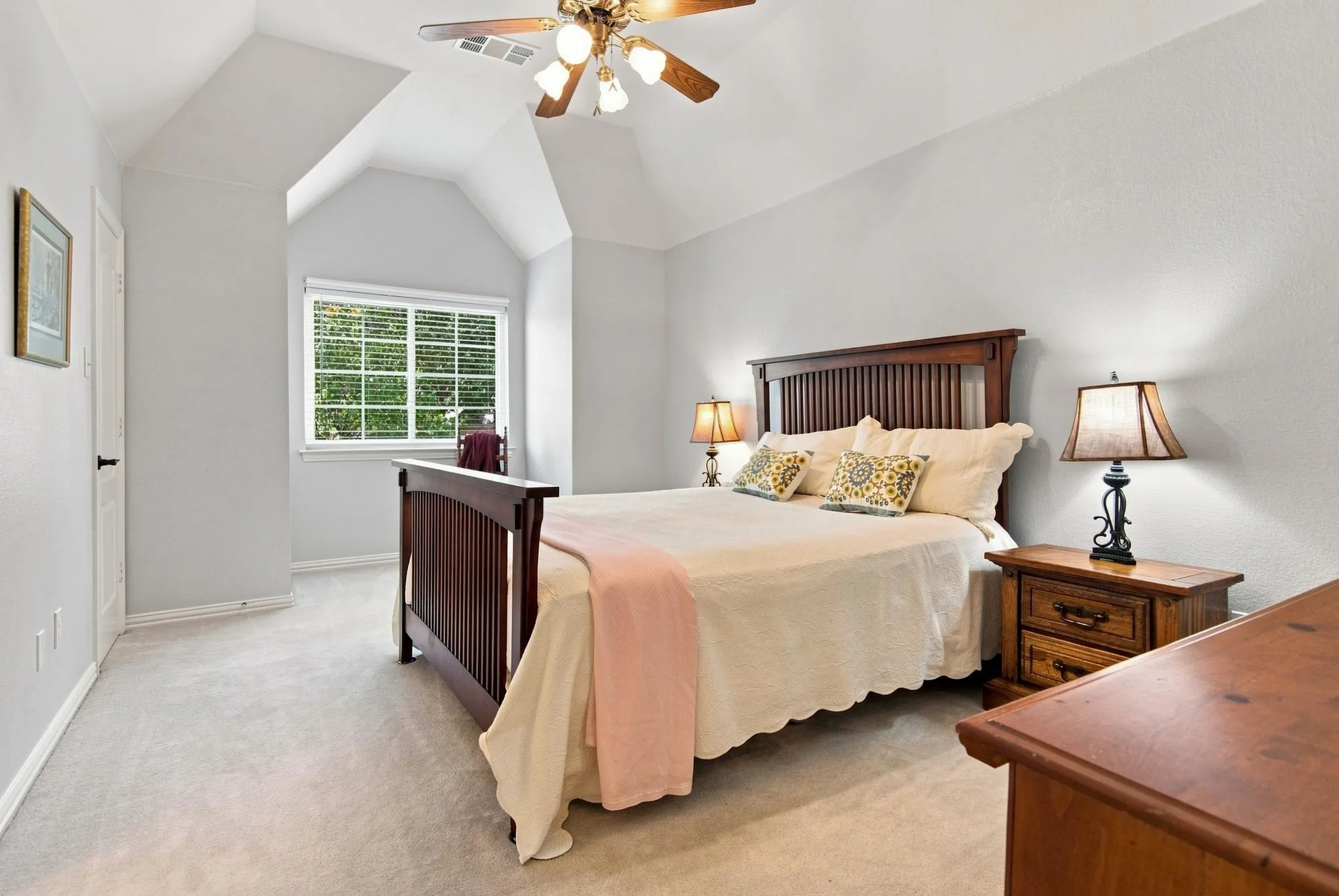 Primary bedroom suite featuring vaulted ceilings with skylight, wood-slat bed frame, carpet flooring, ceiling fan with integrated lighting, paired nightstands with wood construction and attached task lighting, and an arched window with multiple panes overlooking landscaped grounds