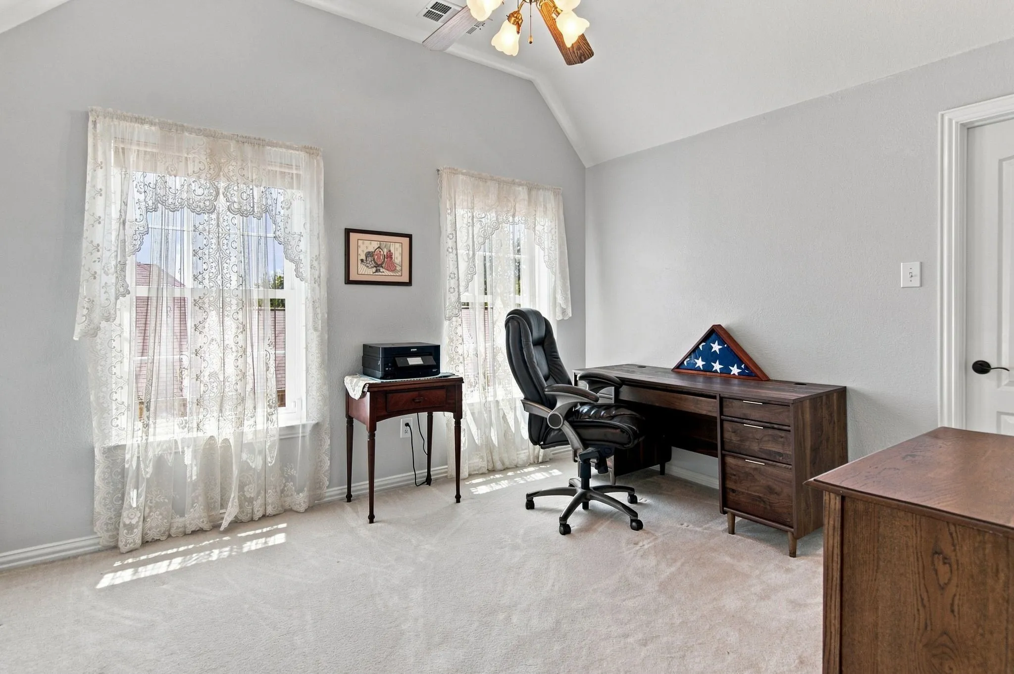 Spacious home office featuring vaulted ceilings with recessed lighting, dual windows with lace curtains providing natural light, carpet flooring, and ample built-in wood cabinetry and desk space for work and storage