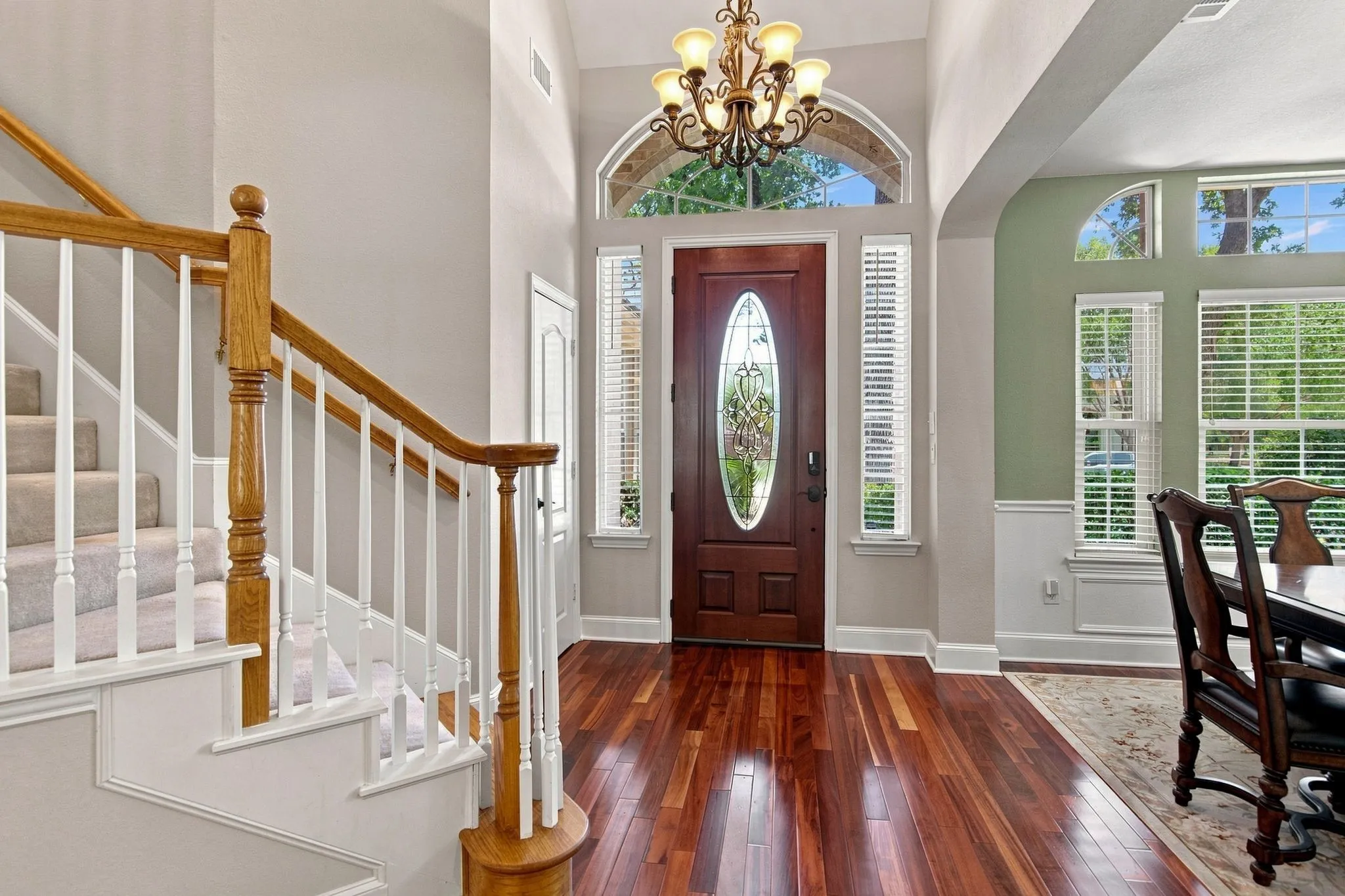 Elegant foyer showcasing rich hardwood flooring, a grand staircase with turned wooden balusters and newel post, soaring ceilings with an arched transom window, a brass chandelier, and a burgundy entry door with sidelights and transom detailing