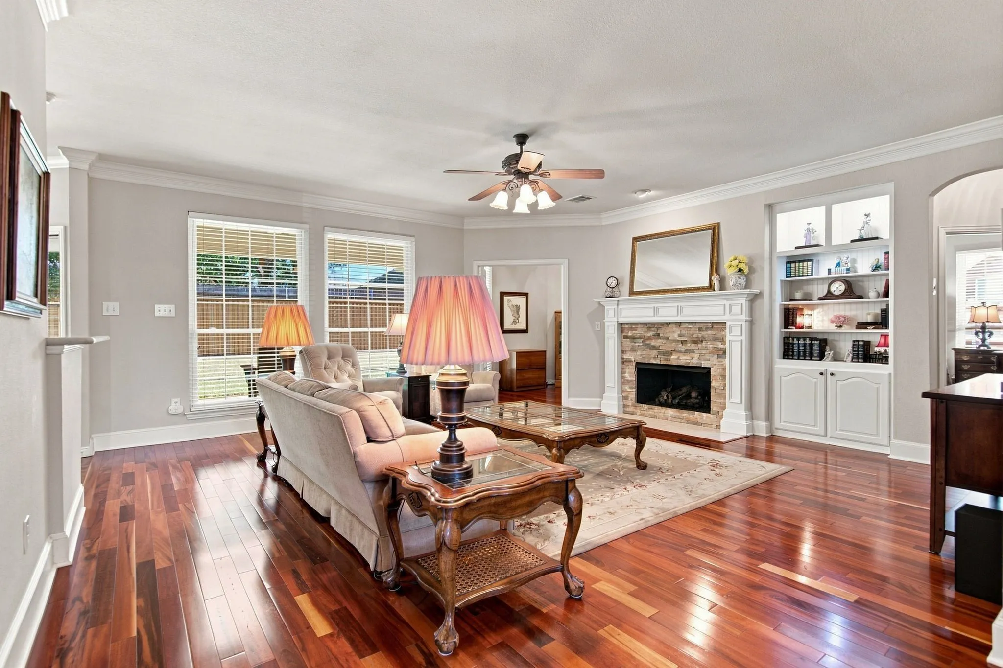Rich wood-finish flooring throughout, decorative fireplace with stone surround, custom built-in shelving, crown molding, and a ceiling fan