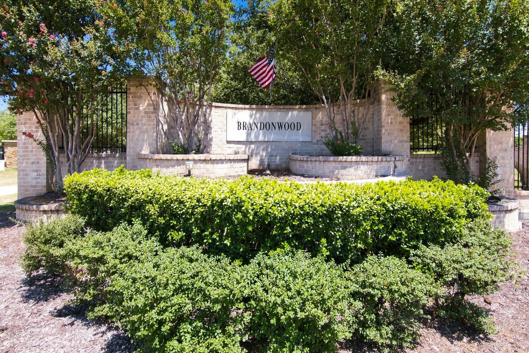 Brandywood community entrance featuring a welcoming brick monument sign, mature shade trees, and manicured landscaping with vibrant green hedges and seasonal plantings