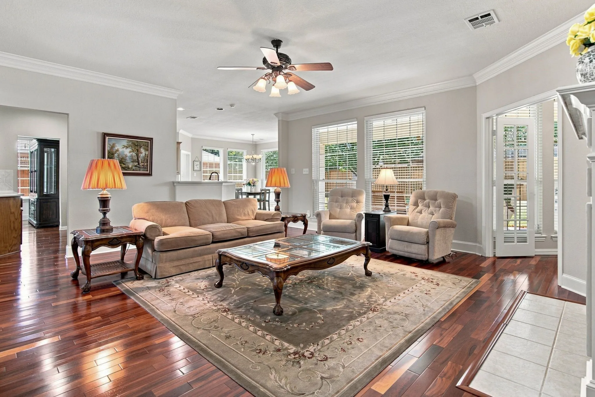 Spacious living room featuring rich wood-finish flooring, ceiling fan with light fixture, and multiple French doors with shutters opening to natural light