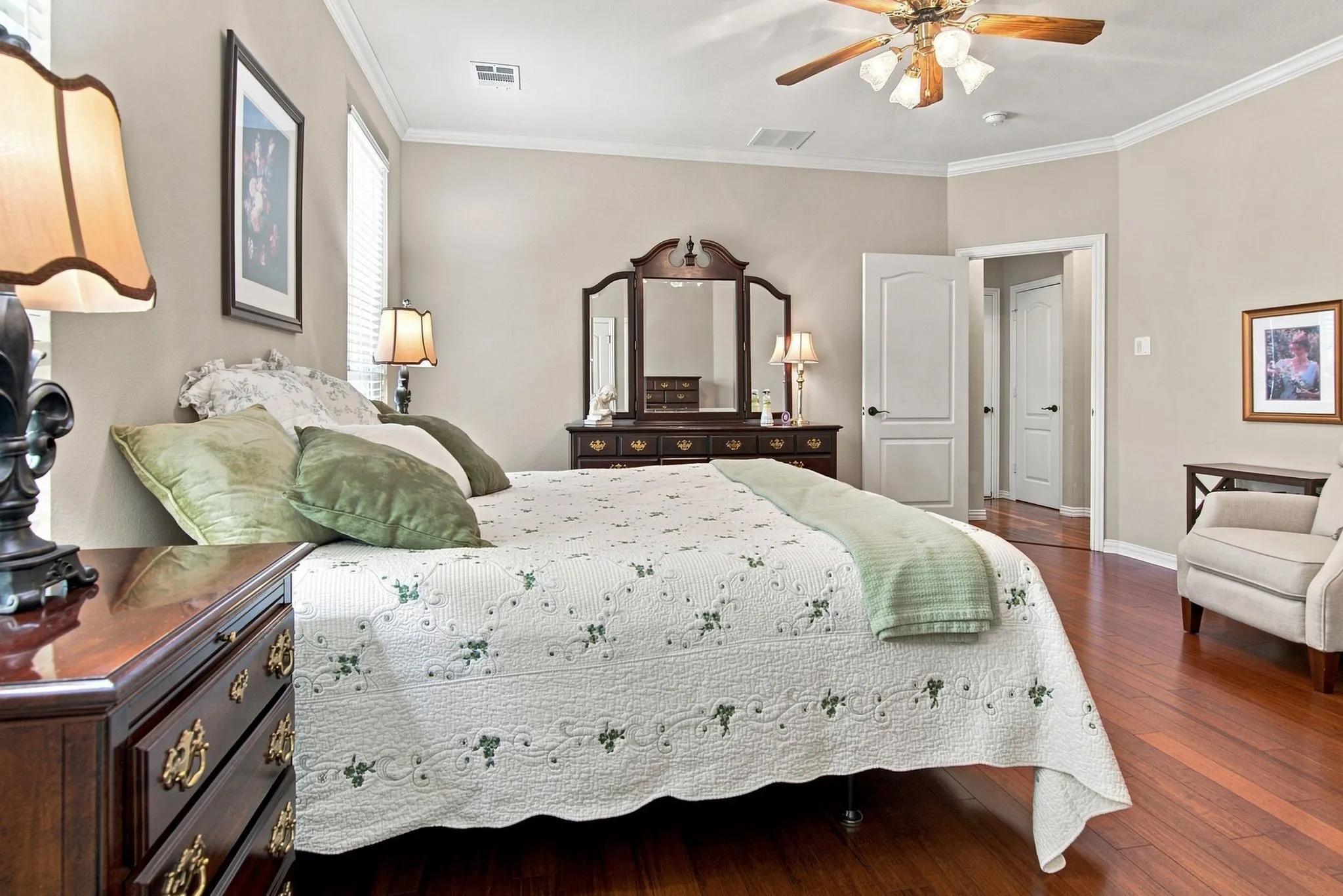 Spacious primary bedroom featuring hardwood flooring, a ceiling fan with integrated lighting, a triple-mirror vanity with attached sconces, and an ensuite bathroom