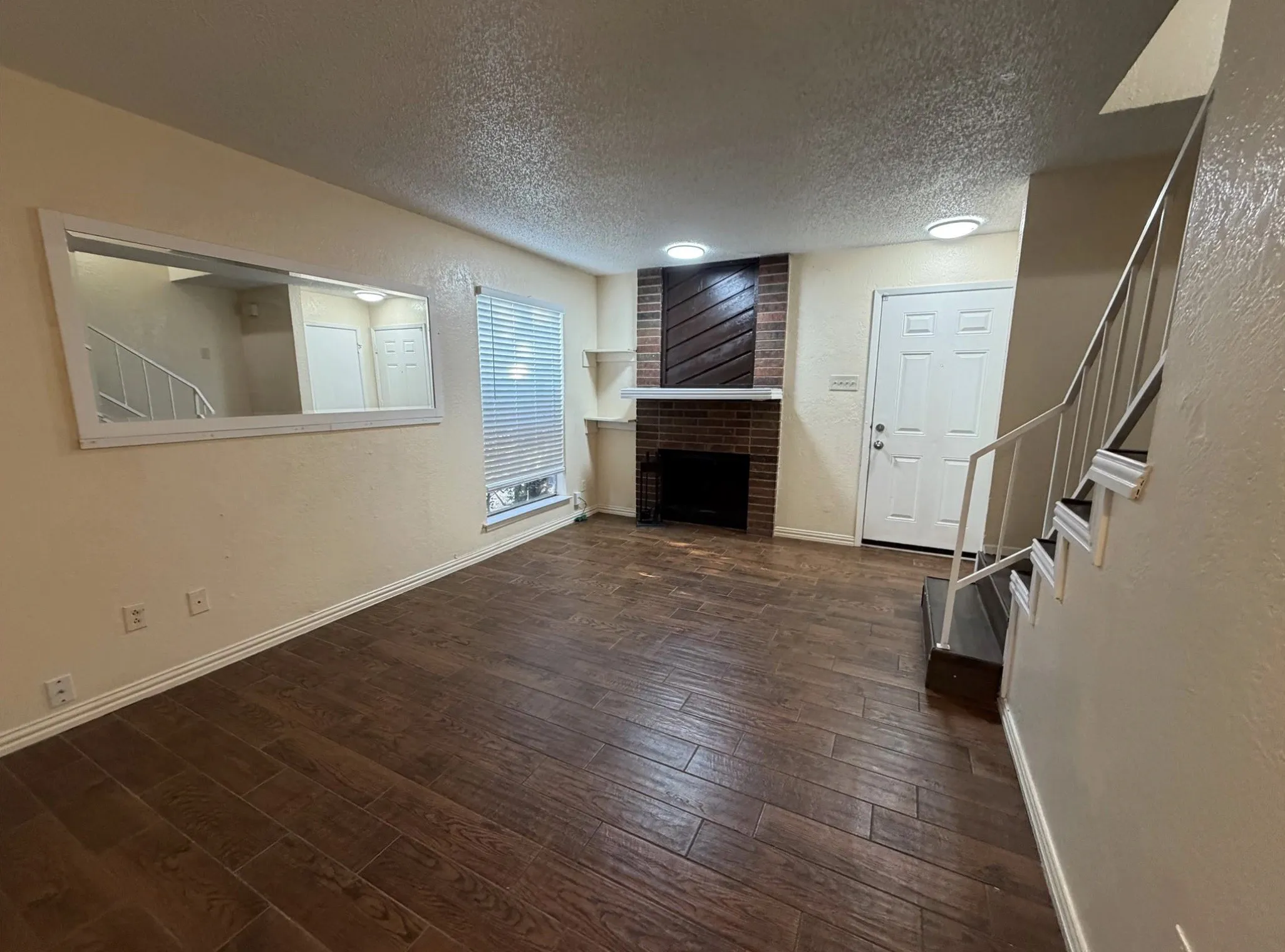 Wood-finish flooring throughout, brick fireplace with mantel, window with blinds, front entry door, and staircase with metal railing