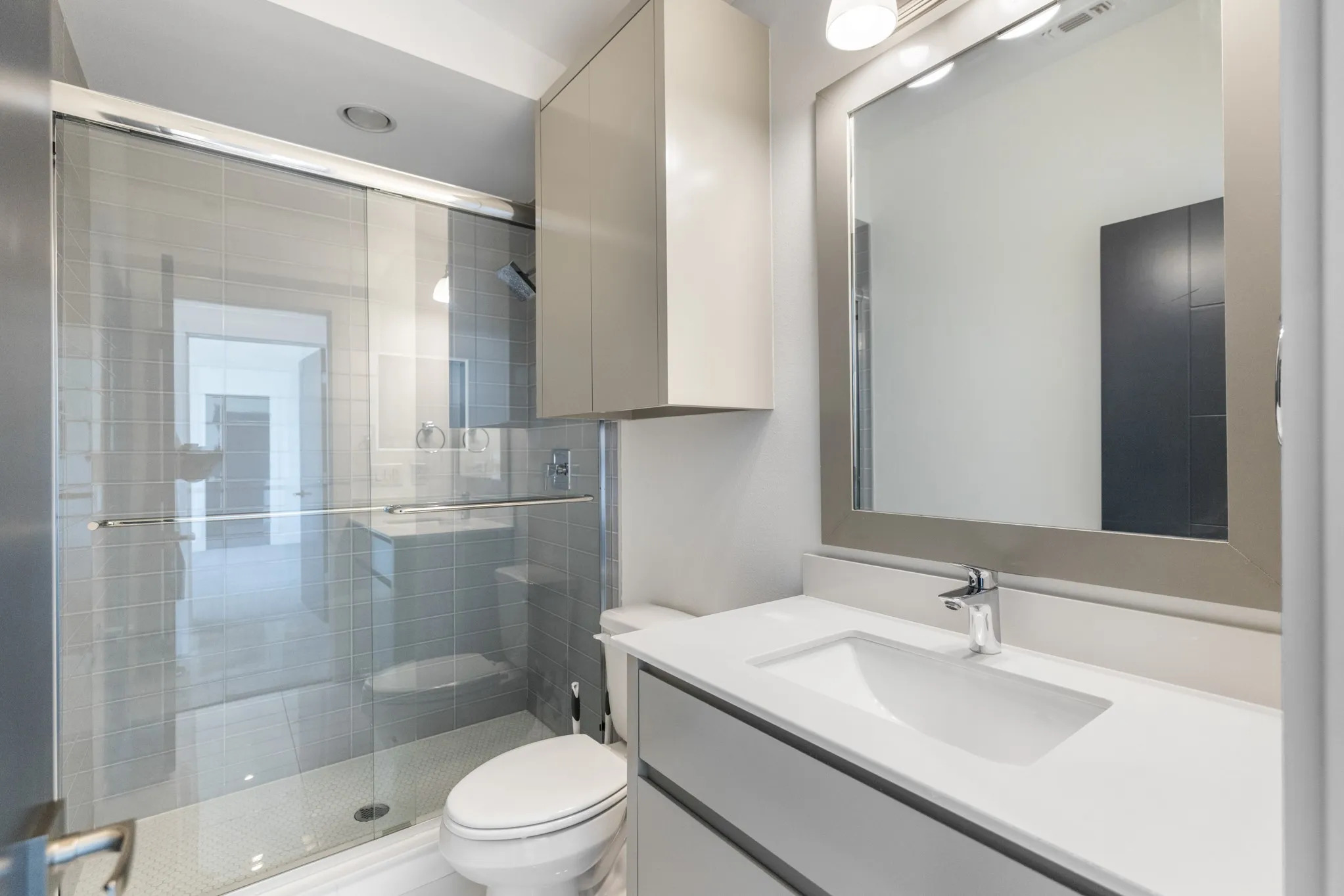 Modern bathroom featuring a frameless glass walk-in shower enclosure with tile surround, a floating vanity with integrated rectangular sink, white quartz countertop, contemporary wall-mounted chrome faucet, and a large framed mirror with integrated lighting