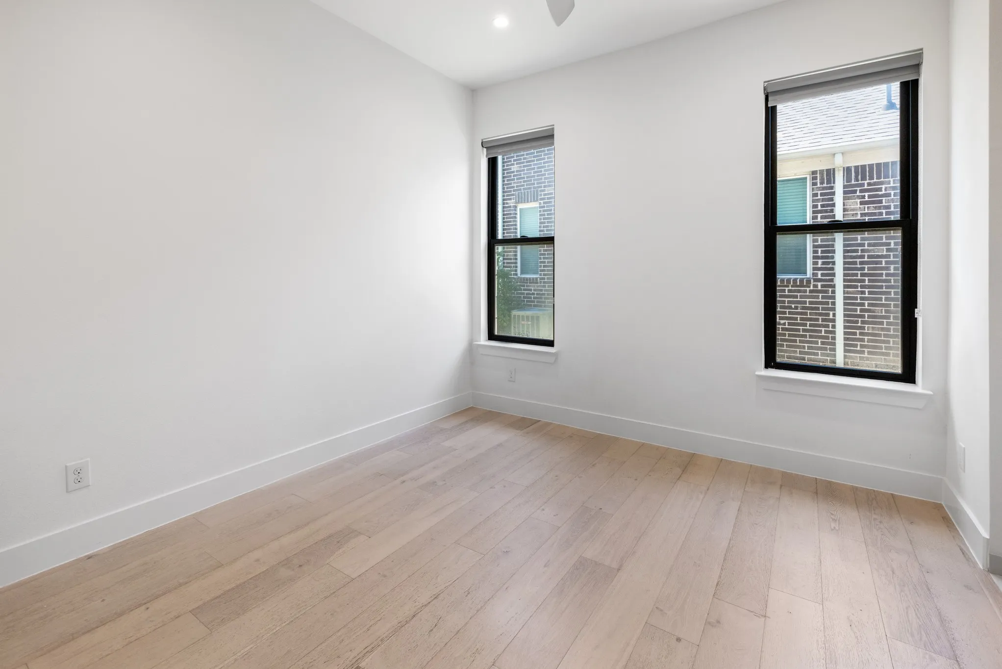 Bright corner bedroom featuring two large double-hung windows with black frames, light wood-finish flooring, and clean white walls with neutral trim