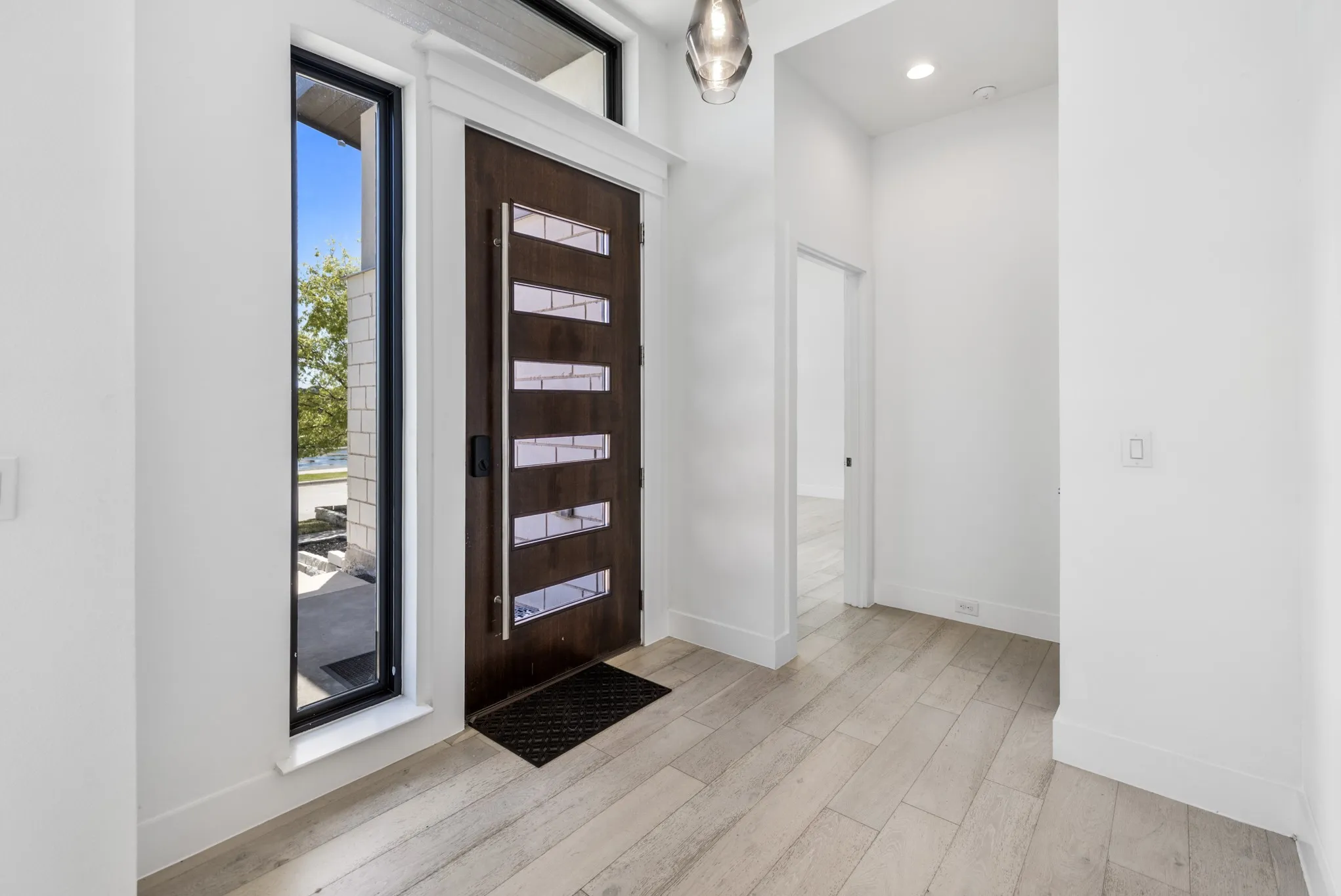 Modern entryway featuring a dark wood contemporary front door with horizontal panel detailing, wide-plank wood-finish flooring, white walls, recessed lighting, and a glass panel entry door with black frame allowing natural light and views of the exterior landscape