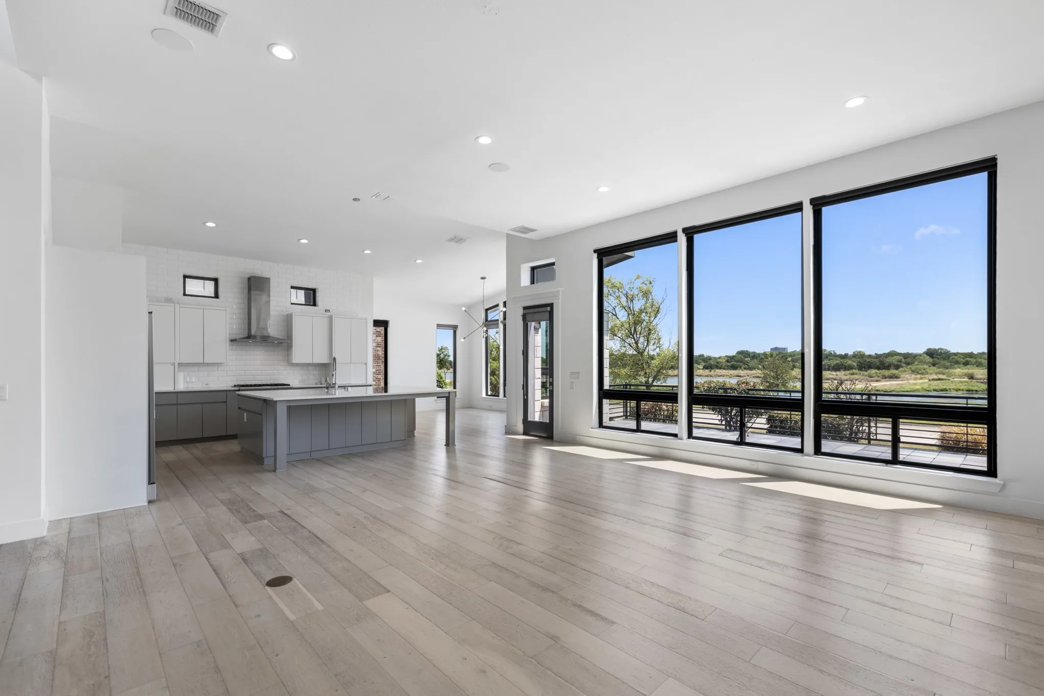 Open-concept living space featuring wide-plank wood-finish flooring, recessed ceiling lighting, and expansive multi-panel glass doors and windows framing pastoral landscape scenery