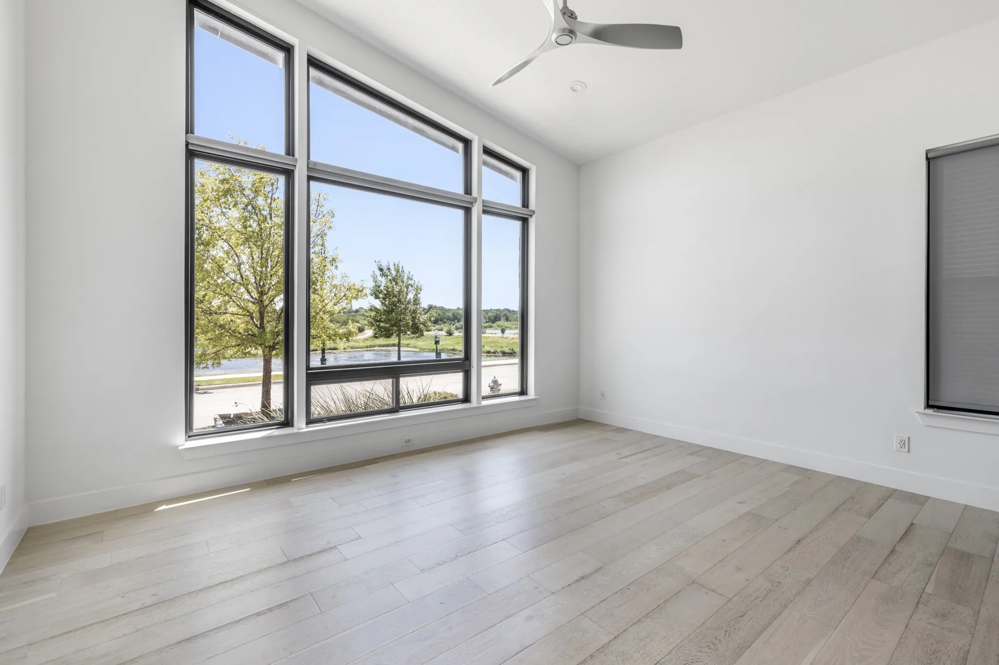 Bright corner room showcasing expansive multi-pane windows with black aluminum frames, light wood-finish flooring, and ceiling fan