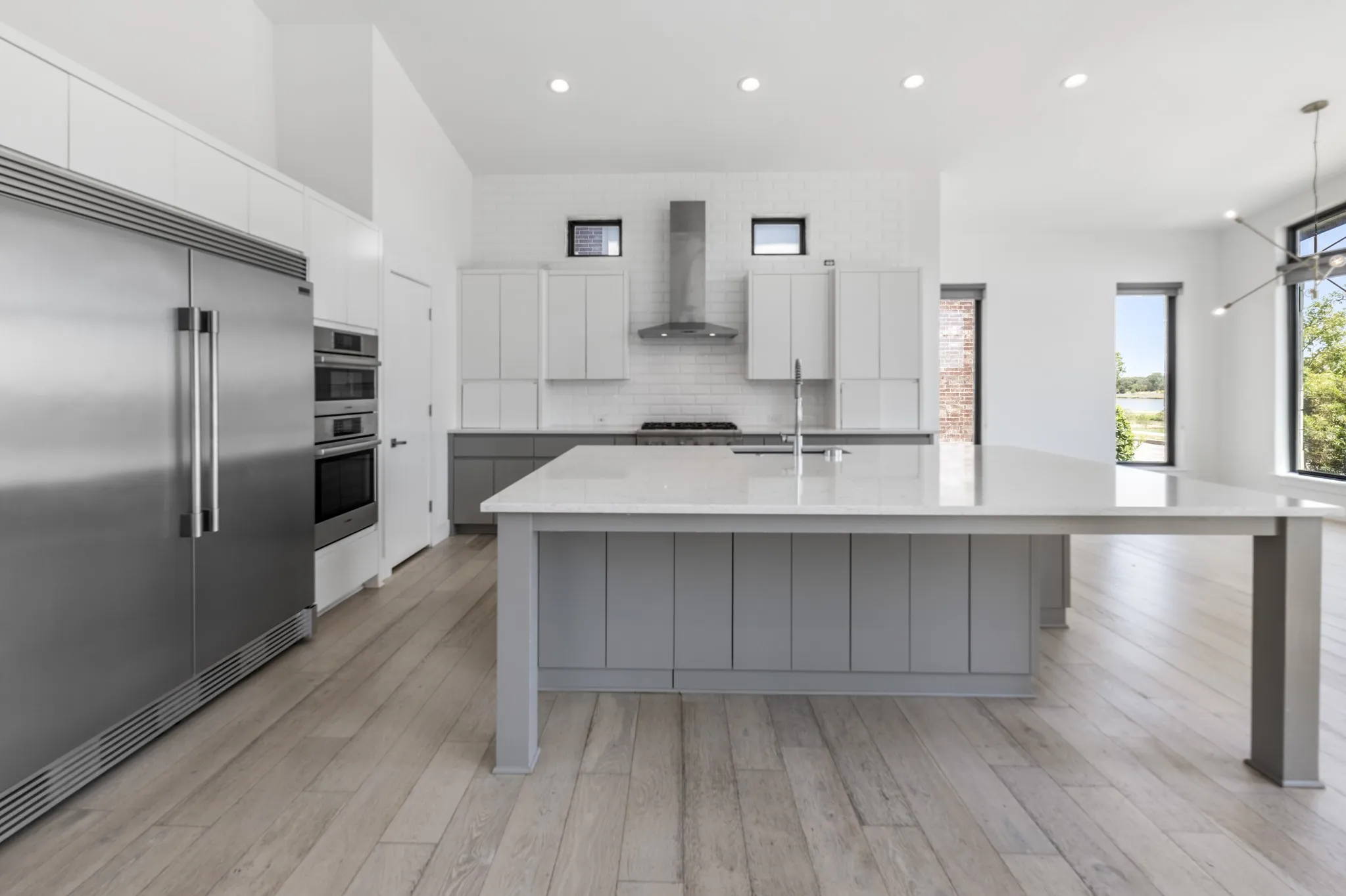 Contemporary kitchen featuring a large waterfall-edge island with gray shiplap paneling, white cabinetry with integrated appliances including stainless steel refrigerator and dual wall ovens, gas cooktop with stainless steel range hood, quartz countertops, and wide-plank wood-finish flooring throughout the open layout