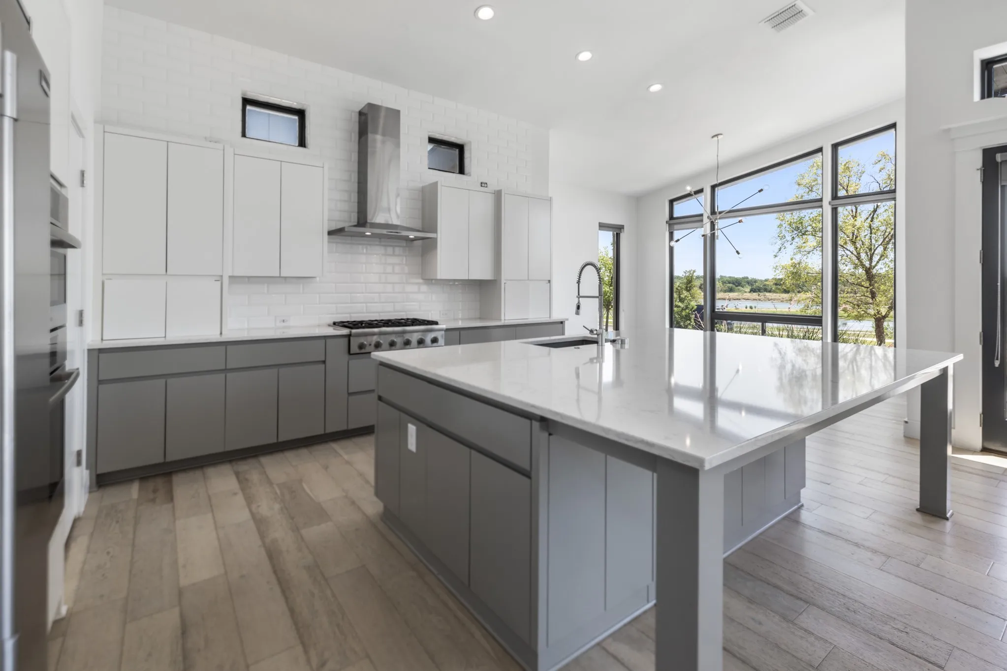 Modern kitchen featuring white upper cabinetry and charcoal lower cabinetry with a large waterfall-edge island, stainless steel range hood, and professional-grade gas cooktop