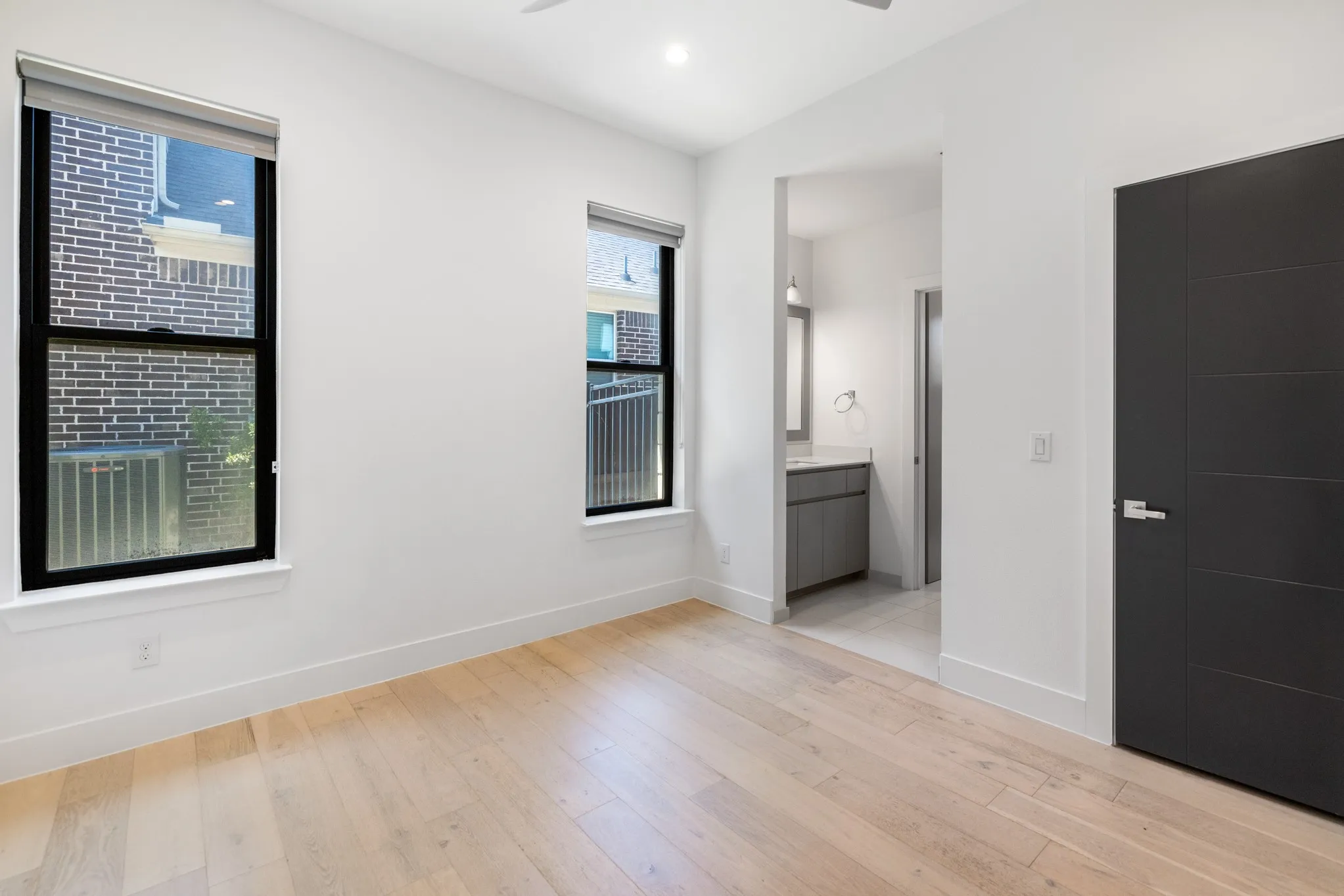 Bright corner bedroom showcasing gleaming wood-finish flooring, abundant natural light from dual windows, and a matte black interior door