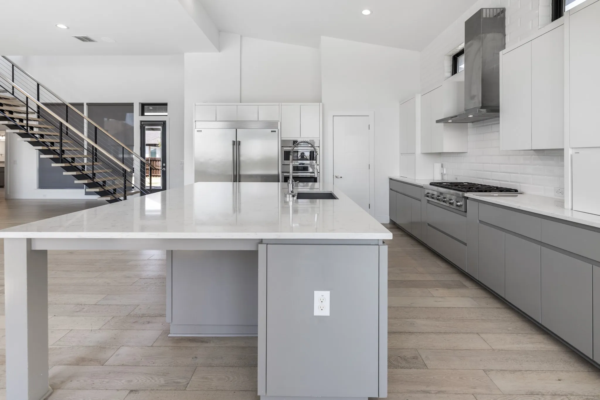 Modern kitchen featuring a large center island with waterfall countertop edge, stainless steel appliances including a built-in refrigerator and range hood, white cabinetry with integrated hardware, quartz countertops, and wood-finish flooring throughout