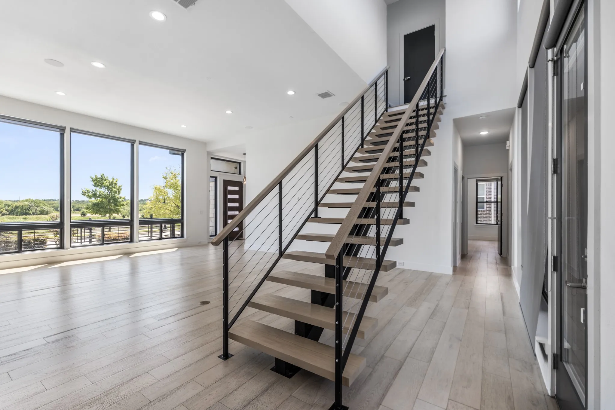Modern open-concept space featuring a floating staircase with black metal railings and wood-finish treads, light wood-finish flooring throughout, floor-to-ceiling windows with expansive natural light, and recessed ceiling lighting