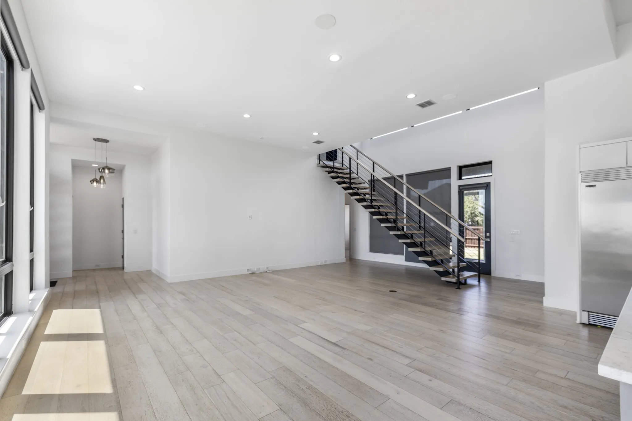 Spacious open-concept interior featuring light wood-finish flooring, high ceilings, recessed lighting, and a modern open-tread staircase with metal railings