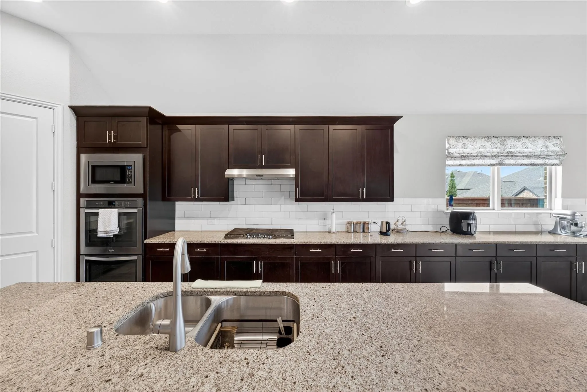 Chef's kitchen featuring dark wood cabinetry with brushed metal hardware, quartz countertops with waterfall island edge, stainless steel dual wall ovens, gas cooktop with subway tile backsplash, and recessed lighting throughout