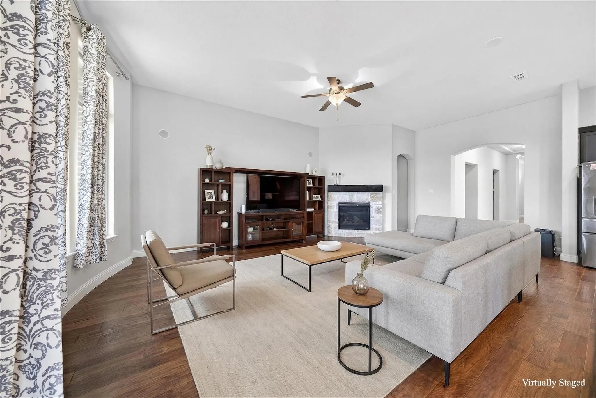 Spacious open-concept living area featuring hardwood flooring, a built-in entertainment center with wood cabinetry, a ceiling fan with brass detailing, and large windows dressed in patterned drapery