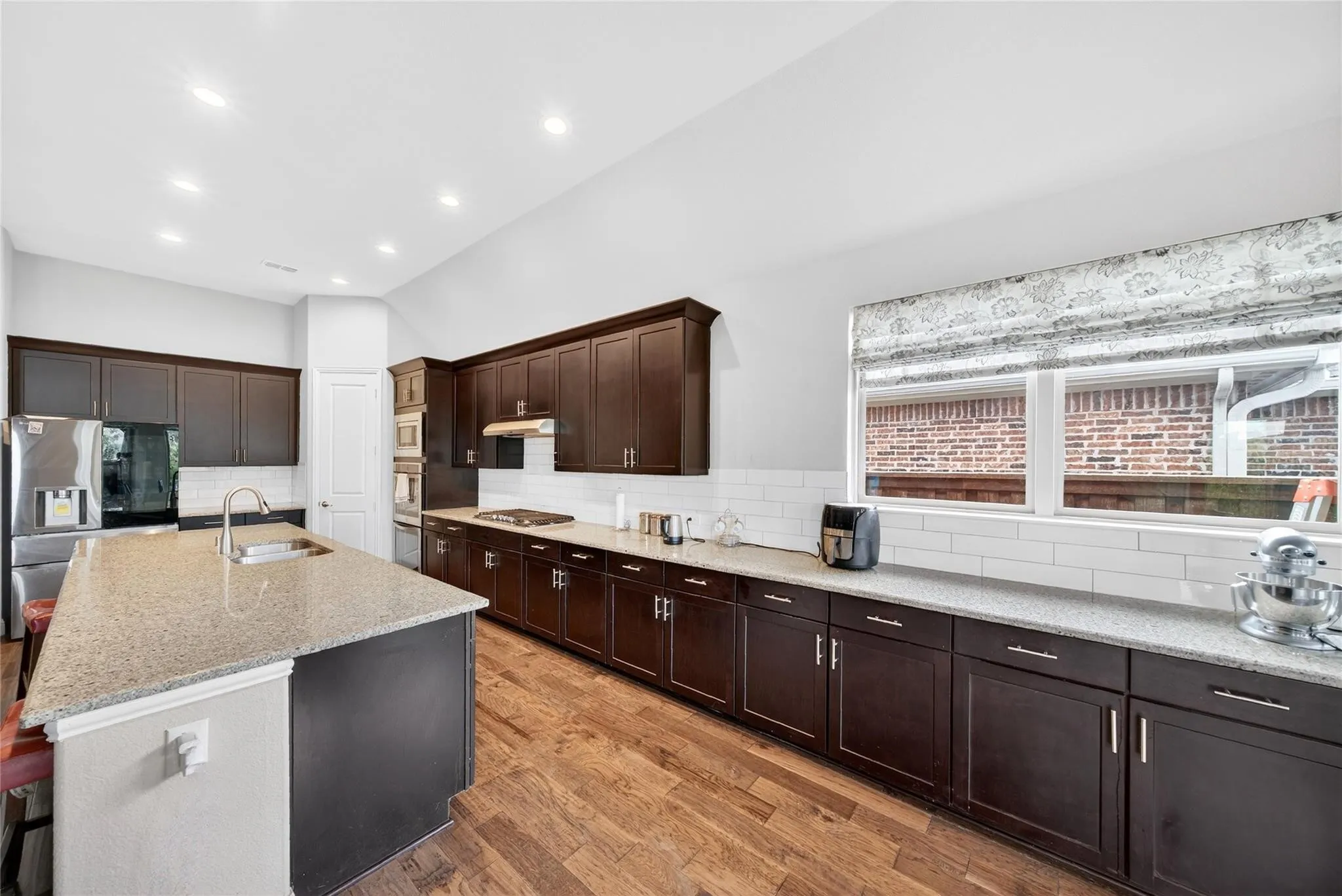 Chef's kitchen featuring dark cabinetry with brushed metal hardware, quartz countertops, a large center island with waterfall edge, stainless steel appliances, subway tile backsplash, recessed lighting, and wood-finish flooring throughout
