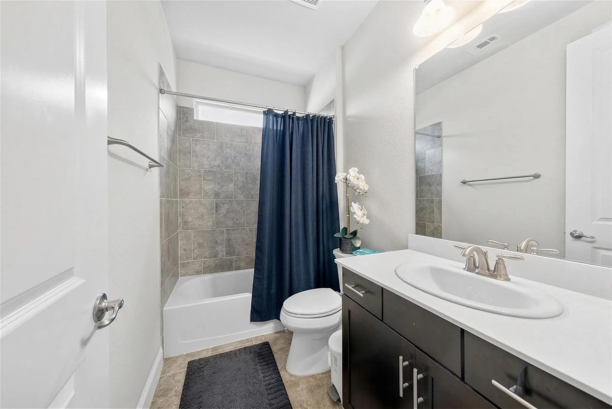 Modern bathroom featuring a dual-sink vanity with dark cabinetry and white quartz countertop, frameless mirror with integrated lighting, a separate soaking tub with tile surround and navy shower curtain, and ceramic tile flooring throughout