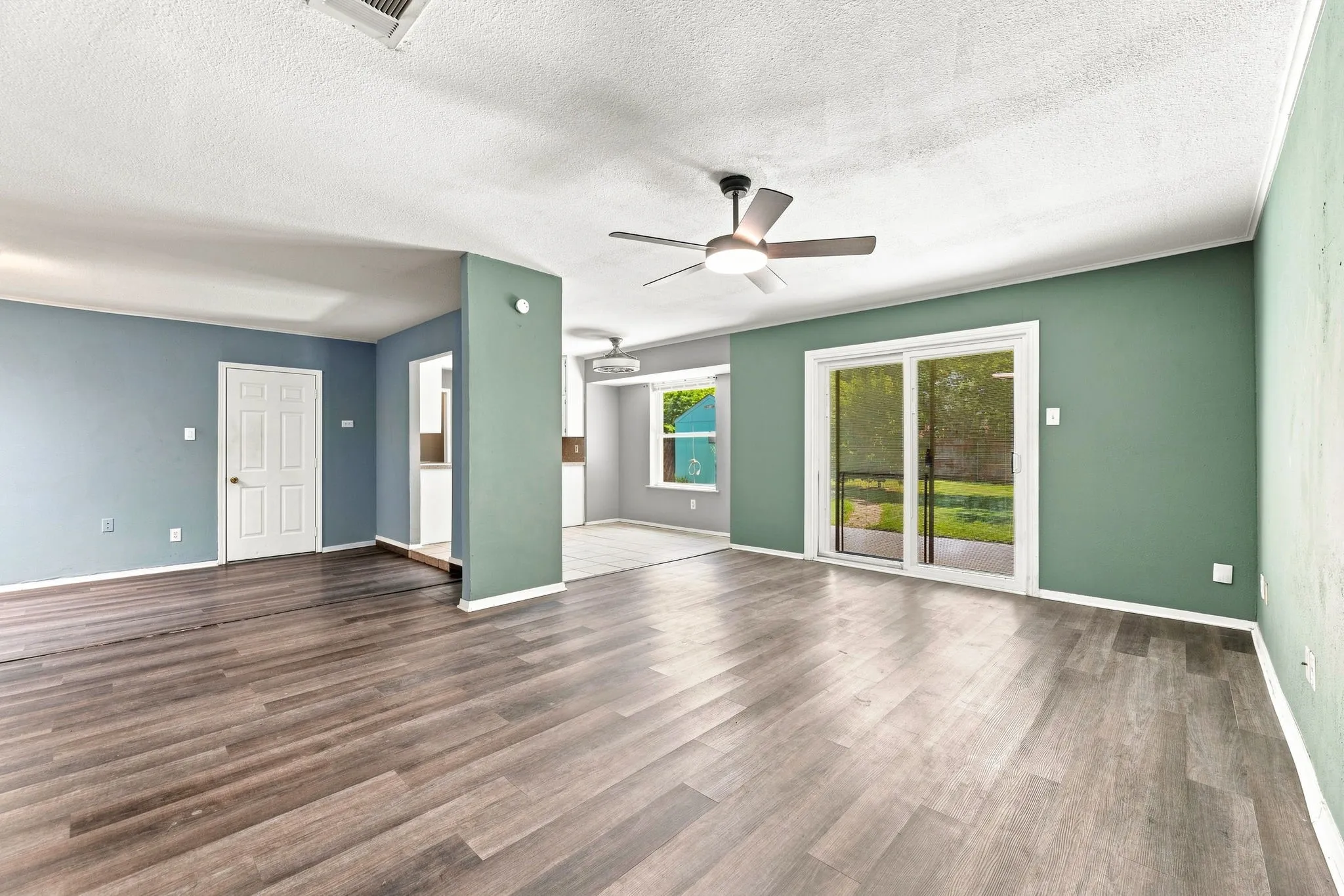 Spacious open-concept living area showcasing wood-finish flooring throughout, sage green accent walls with white trim, large glass sliding doors with natural light exposure, and a ceiling fan with brushed metal finish