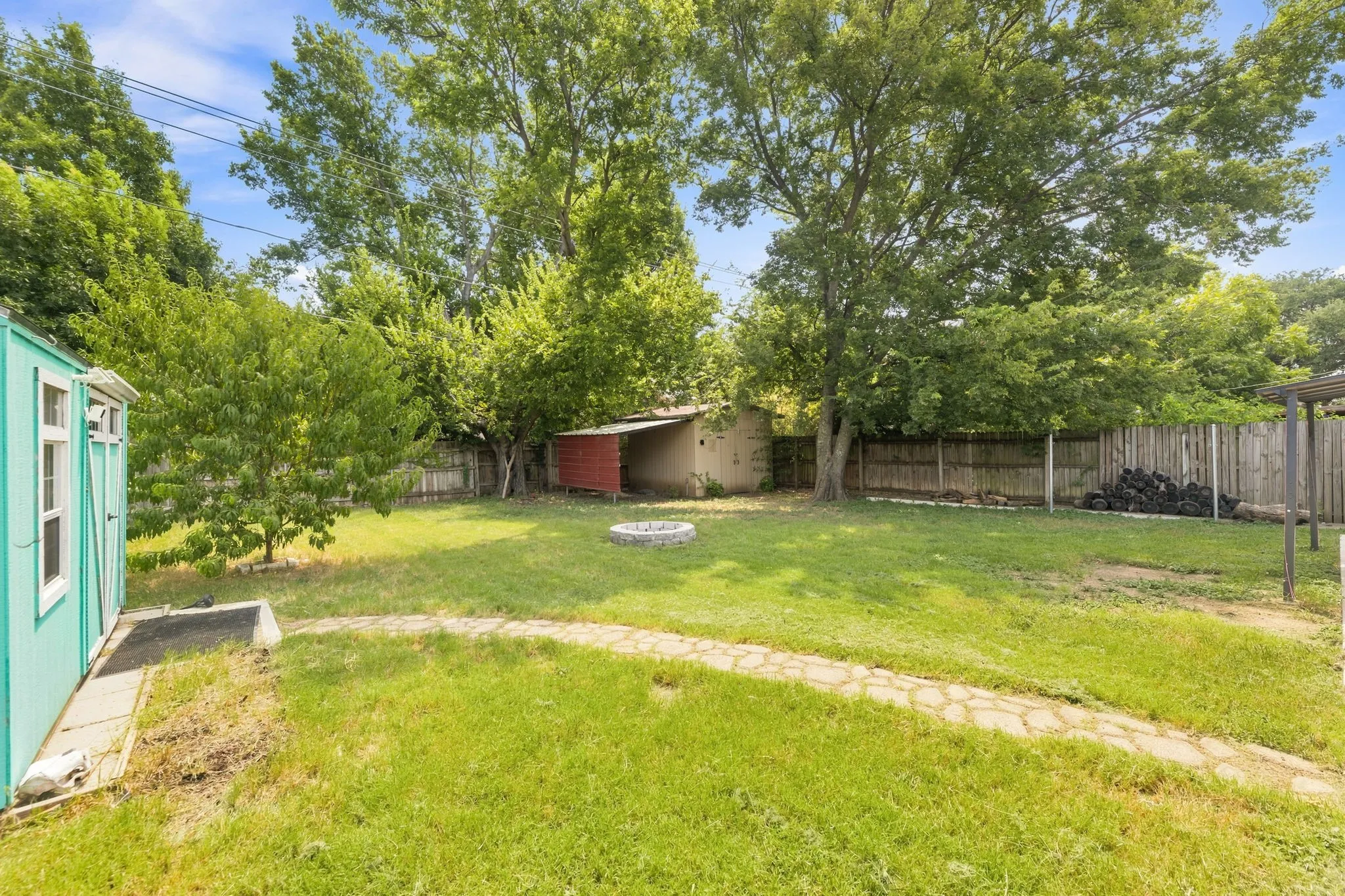 Expansive backyard featuring a mature tree canopy, level lawn, wooden fencing, a detached storage structure, and a fire pit area—ideal for outdoor entertaining and recreation