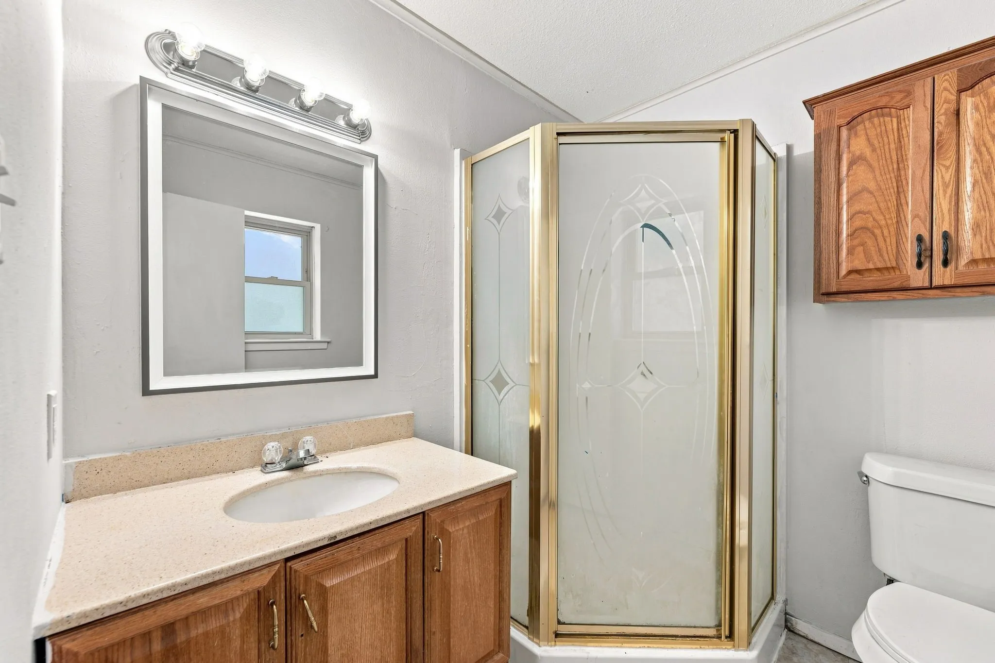 Spa-inspired bathroom featuring a wood-finish vanity with dual-sink configuration and light stone countertop, illuminated framed mirror with integrated lighting, frameless glass shower enclosure with brass-tone hardware and diamond-pattern glazing, and white ceramic toilet