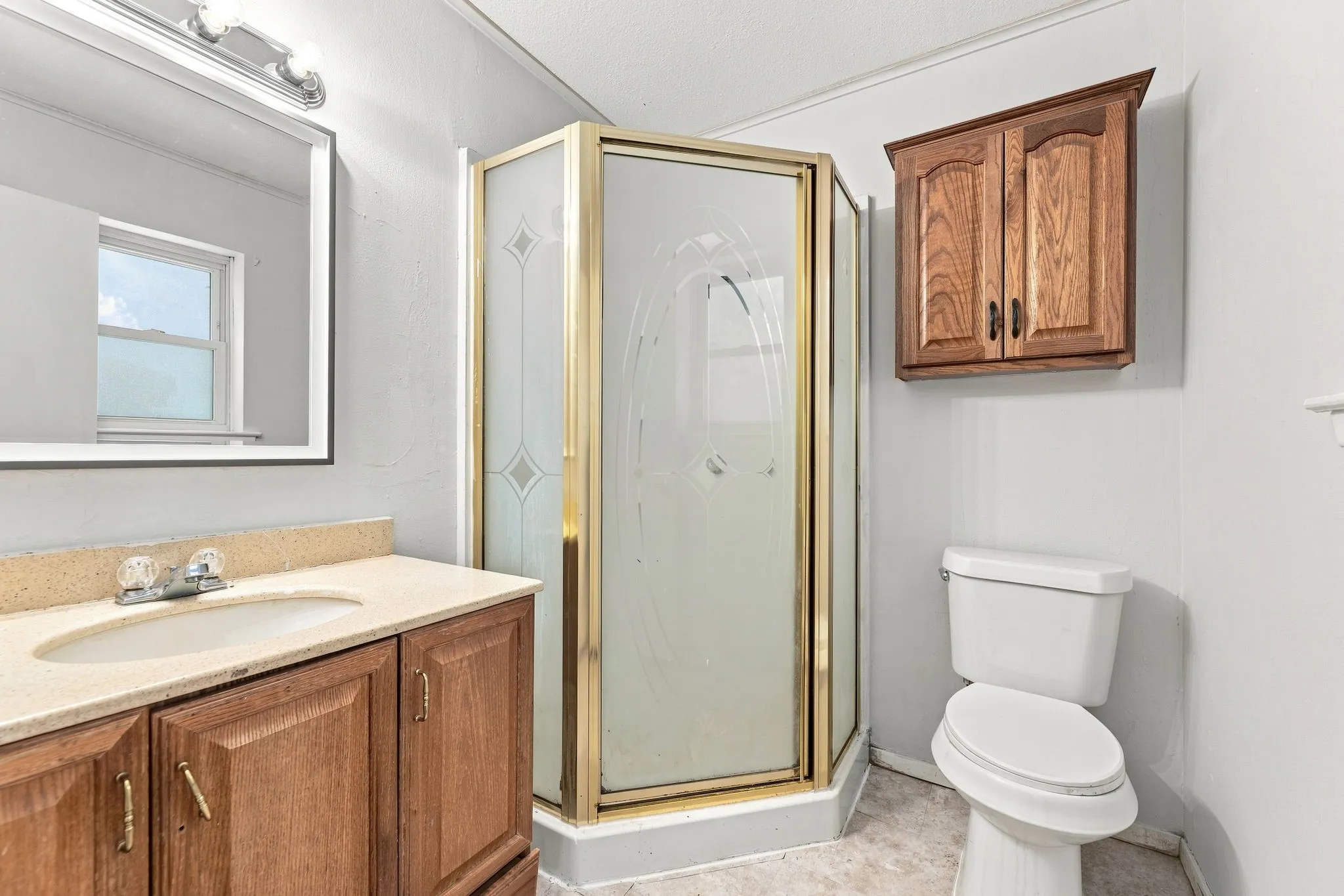 Spa-inspired bathroom featuring dual vanity with wood cabinetry, stone countertops, framed mirror with integrated lighting, corner shower enclosure with brass-tone framing and frosted glass panels, and ceramic tile flooring
