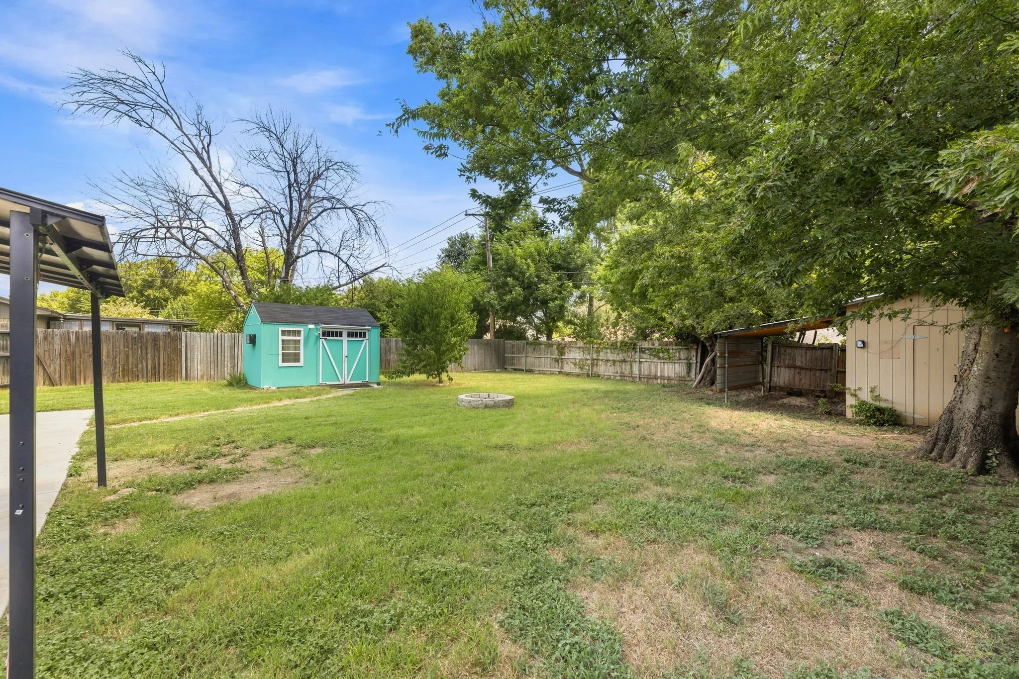 Expansive backyard featuring a mature tree-lined perimeter, level lawn, detached storage shed with turquoise trim, fire pit area, and wooden pergola structure along the left side