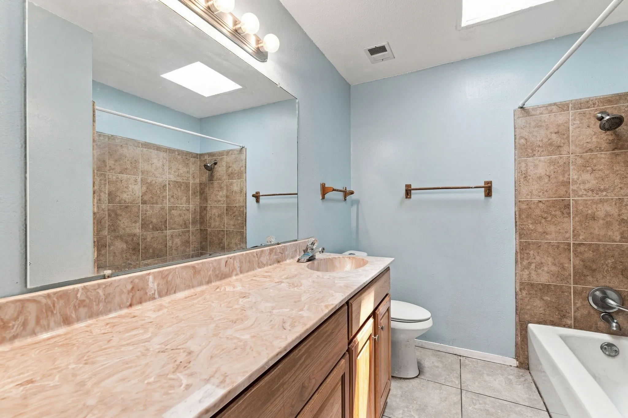 Bathroom featuring a wide vanity with integrated sink, wood-finish cabinetry, a large wall-mounted mirror, tiled shower surround, and a ceiling-mounted skylight