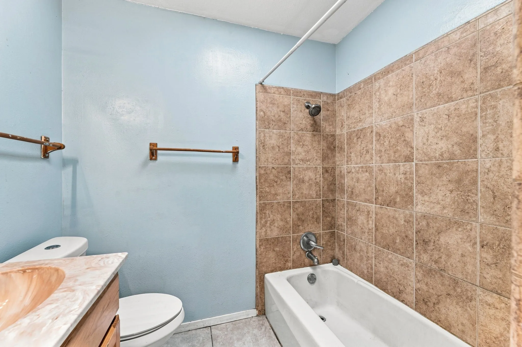 Updated bathroom featuring light blue painted walls, ceramic tile flooring, wood-finish vanity with integrated sink, toilet, and a soaking tub with tile surround and chrome fixtures