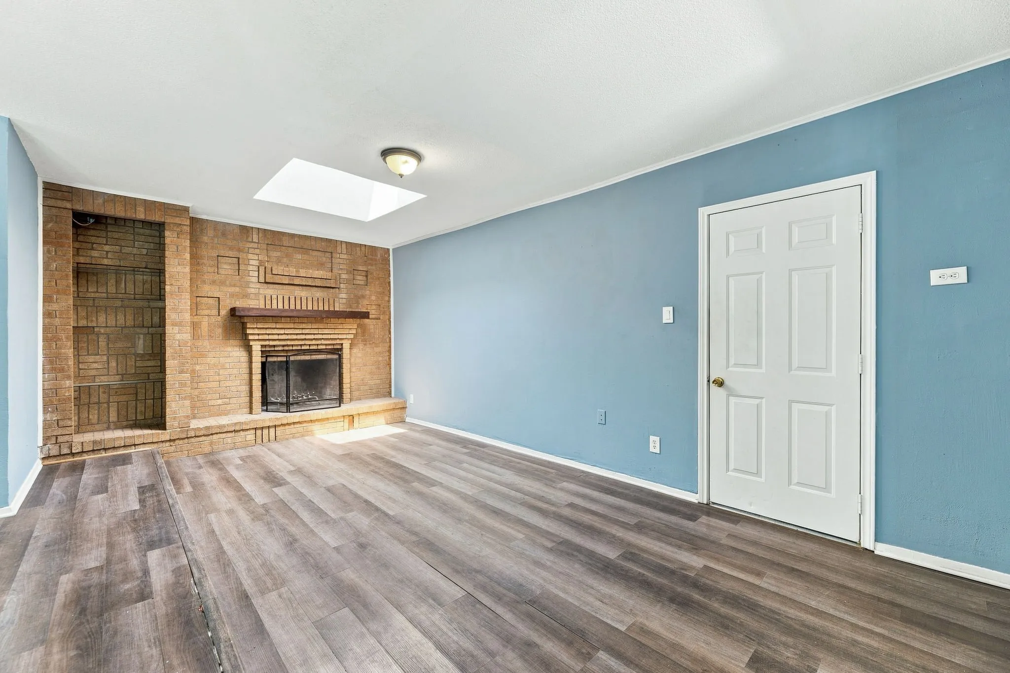 Brick fireplace with a wood mantel, skylight, recessed brick shelving, and wood-finish flooring