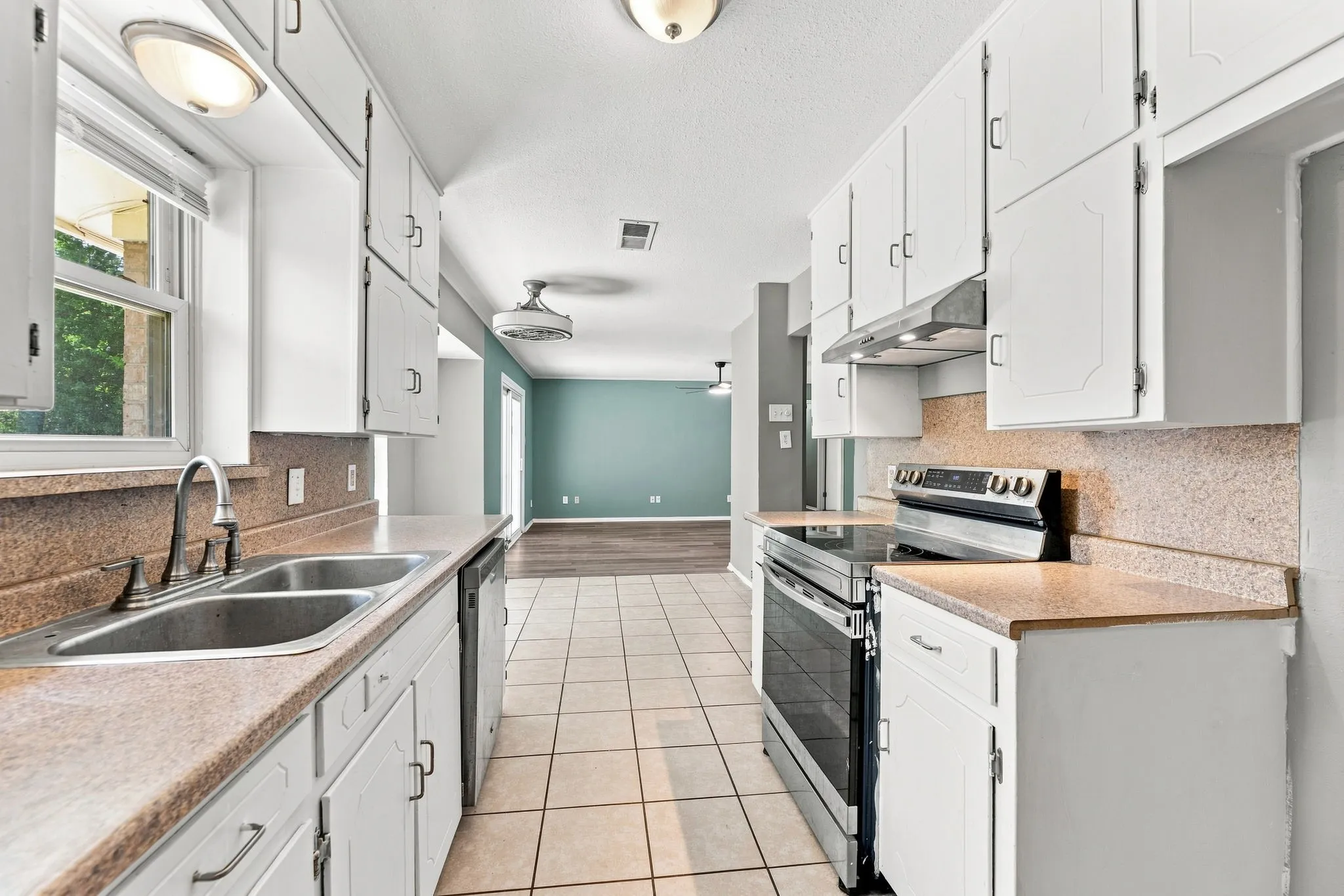 Galley kitchen featuring white cabinetry with hardware accents, wood-finish countertops, ceramic tile flooring, stainless steel appliances including a gas range, undermount sink, and abundant natural light from a nearby window