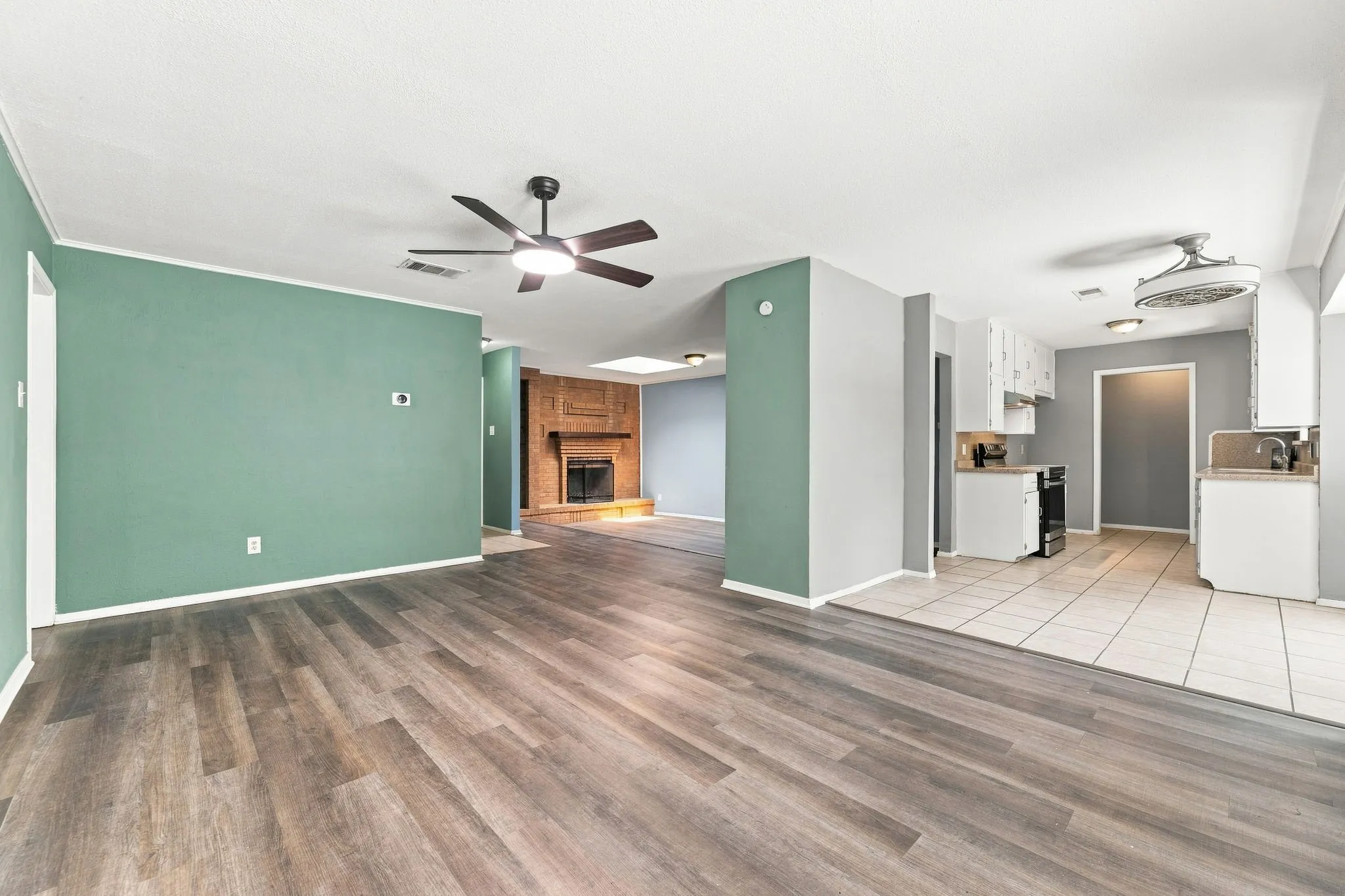 Wood-finish flooring, ceiling fan with integrated lighting, brick fireplace, white cabinetry, and light-colored tile flooring