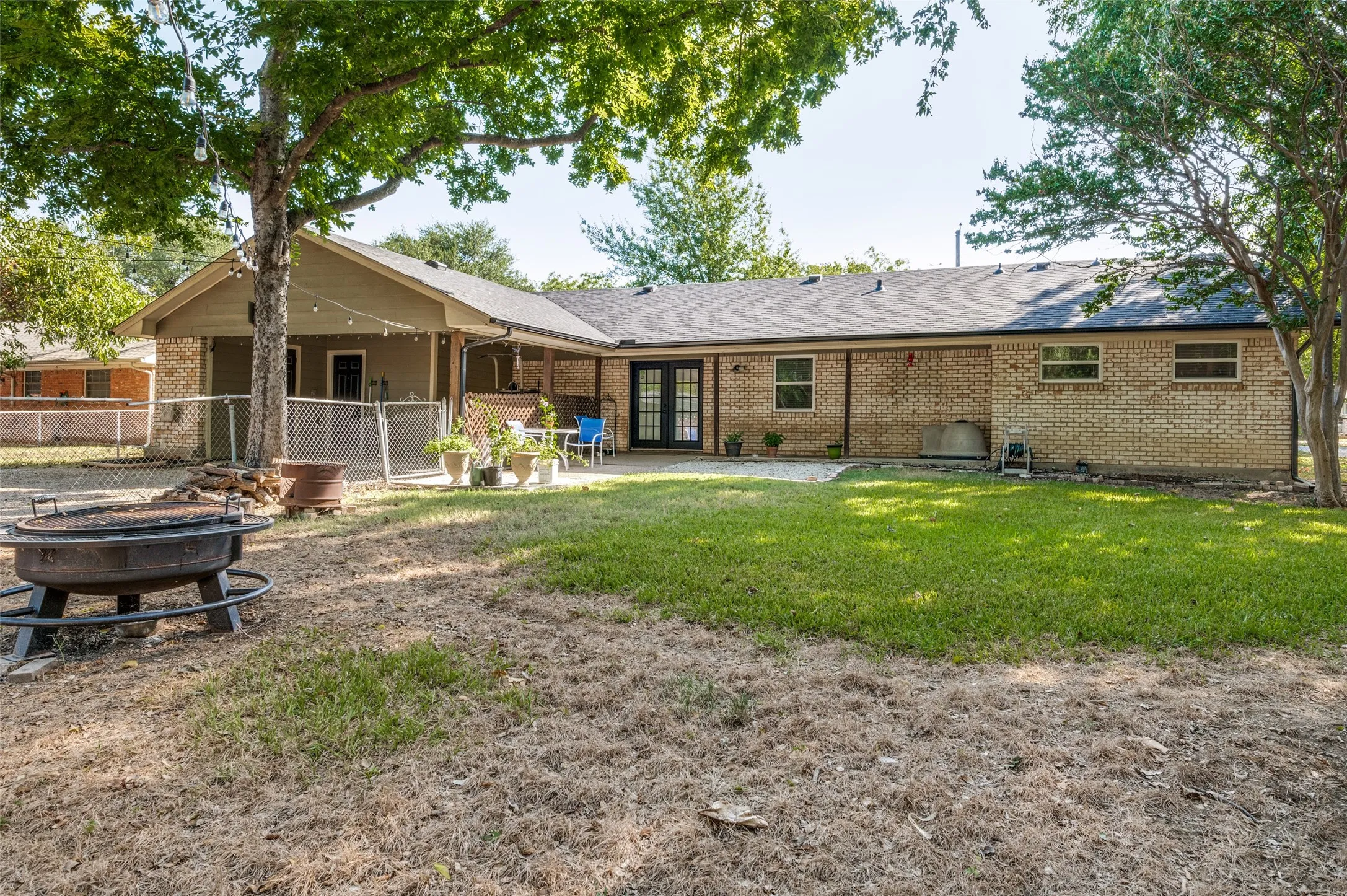 Expansive backyard oasis featuring mature shade trees, manicured lawn, and a fire pit area, with a brick single-story home offering covered patio space and multiple outdoor entertaining zones