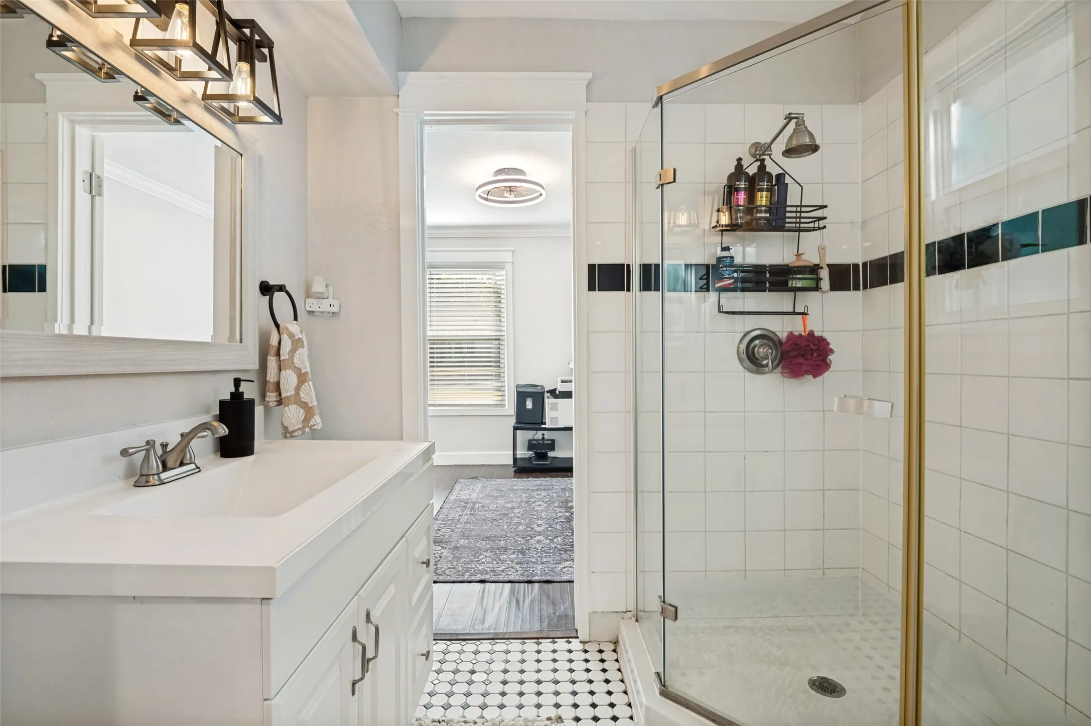 Spa-inspired primary bathroom featuring a walk-in shower with subway tile surround and brass-finished fixtures, dual vanity with white cabinetry and integrated sink, black and white geometric floor tile, and a separate water closet