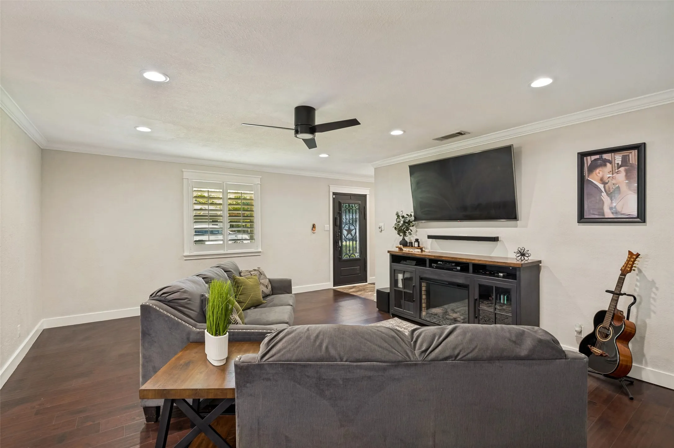 Spacious entertainment area featuring hardwood flooring, a ceiling fan with recessed lighting, a wall-mounted flat-screen television above a dark wood media console with open shelving, and shuttered windows providing natural light