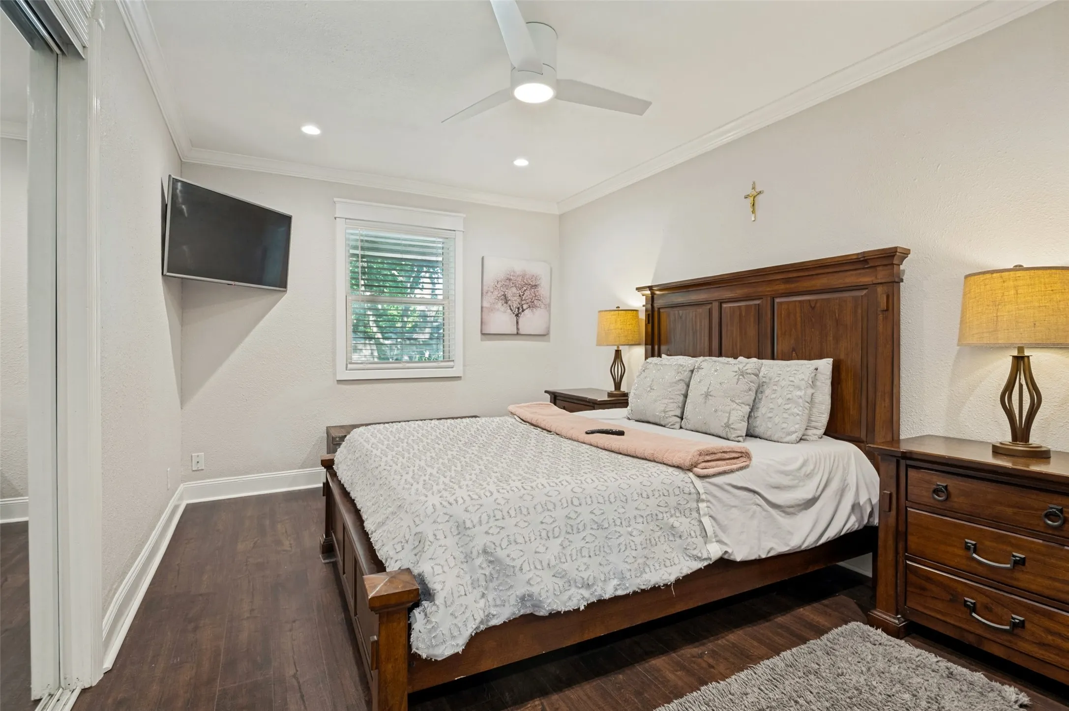 Serene primary bedroom featuring a wood-frame bed with upholstered headboard, hard wood flooring, recessed and ceiling fan lighting, wall-mounted flat-screen television, wood-finish nightstands with brass hardware, and soft neutral wall tones accented by decorative cross and framed artwork