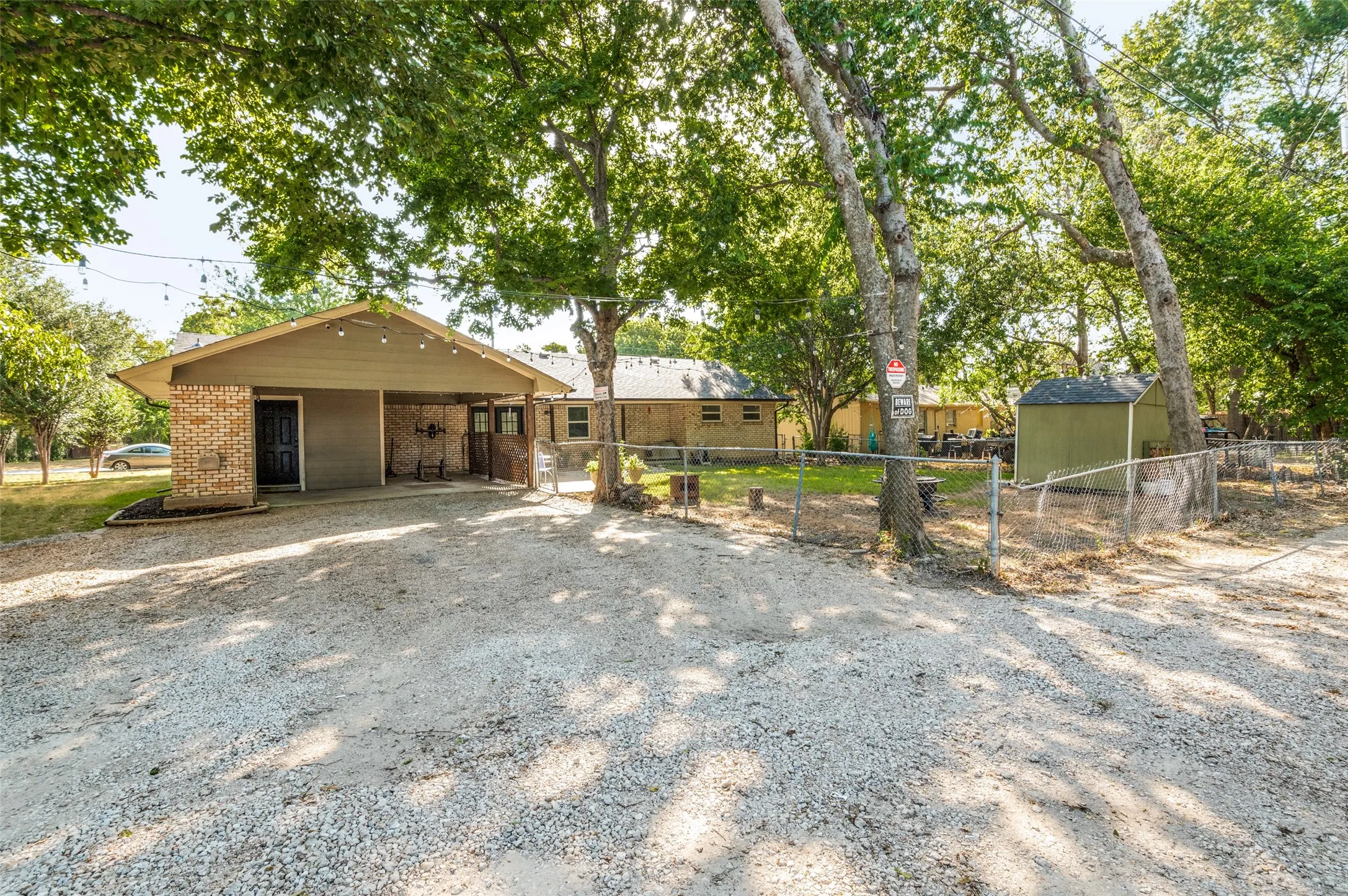 Gravel driveway, covered carport, brick exterior accents, chain-link fencing, mature trees