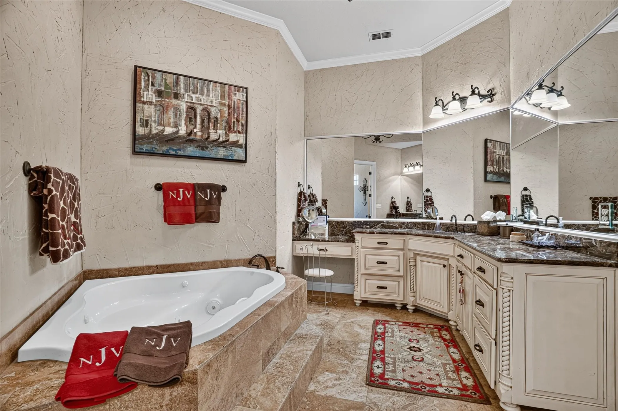 Luxurious primary bathroom featuring a soaking tub with tile surround, dual-sink vanity with cream cabinetry and decorative hardware, stone countertops, and large mirror with integrated lighting