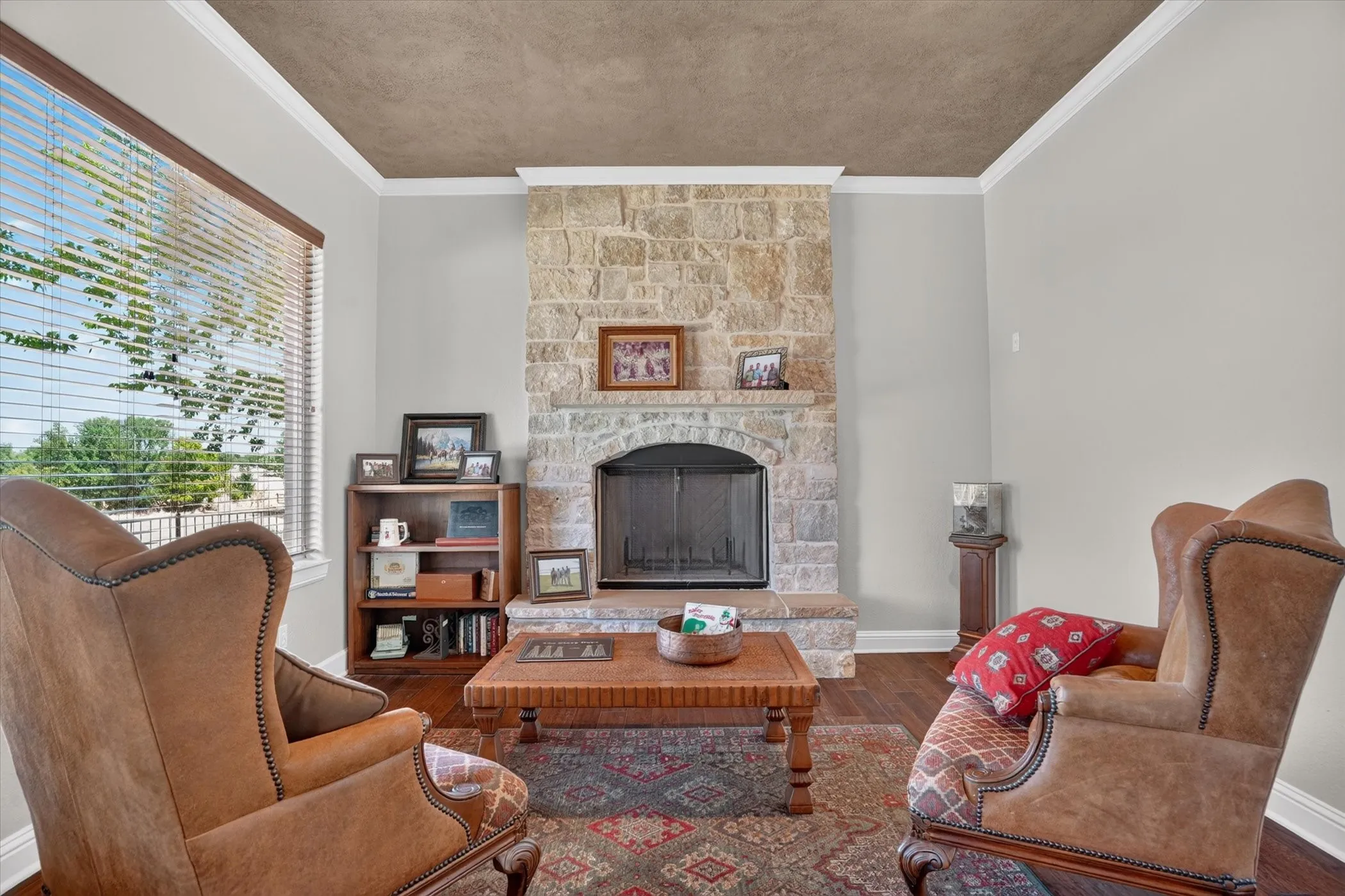 Elegant living room showcasing a stone fireplace with dark metal surround and mantel display, paired wingback chairs with cognac leather upholstery, hardwood-finish flooring, geometric area rug, wooden coffee table, built-in shelving unit with display space, and coffered ceiling detail with neutral wall finishes