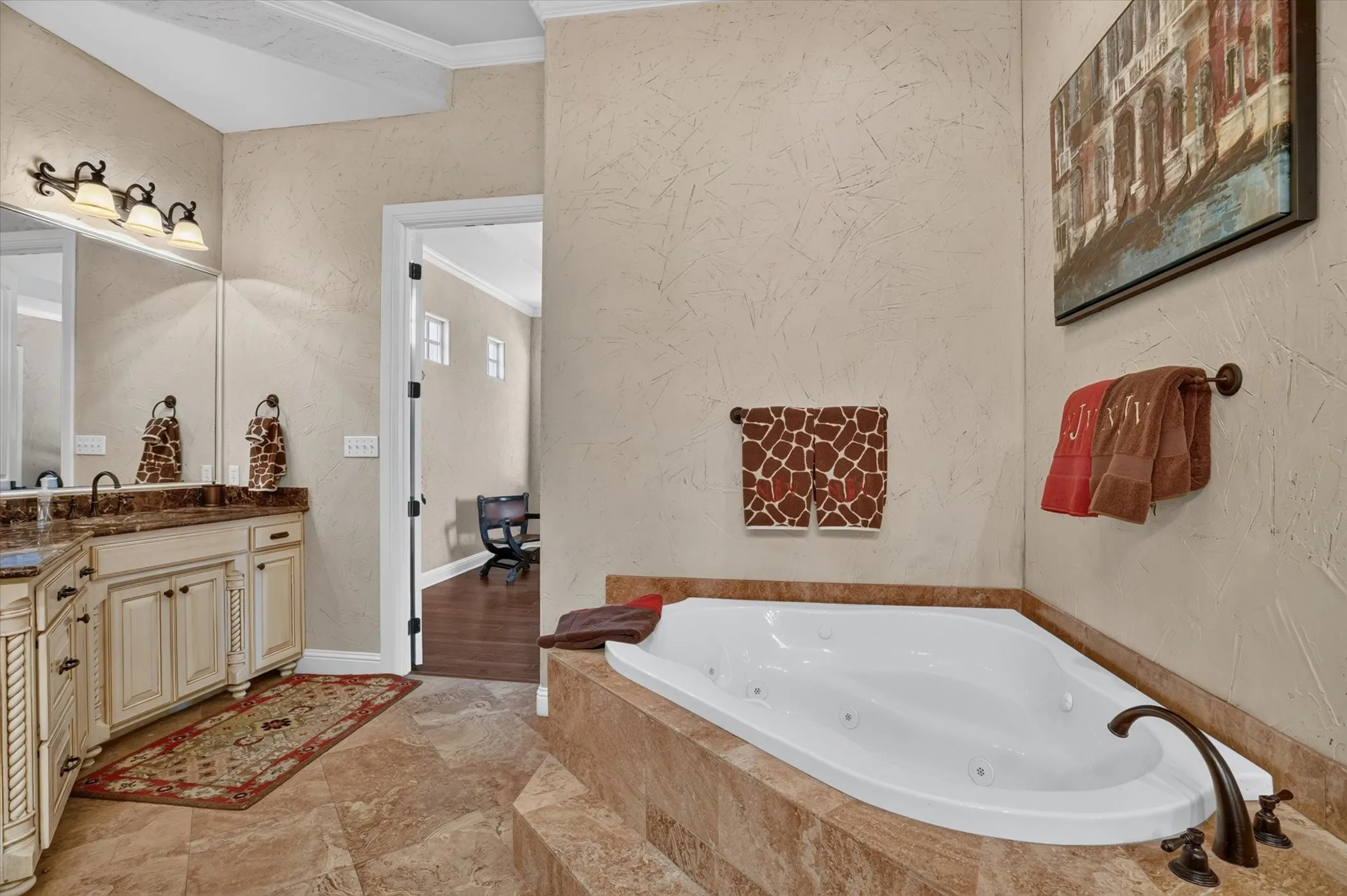 Luxurious spa-inspired master bath featuring a corner soaking tub with integrated jets, dual-sink vanity with ample storage, travertine-look tile flooring, and warm neutral finishes throughout