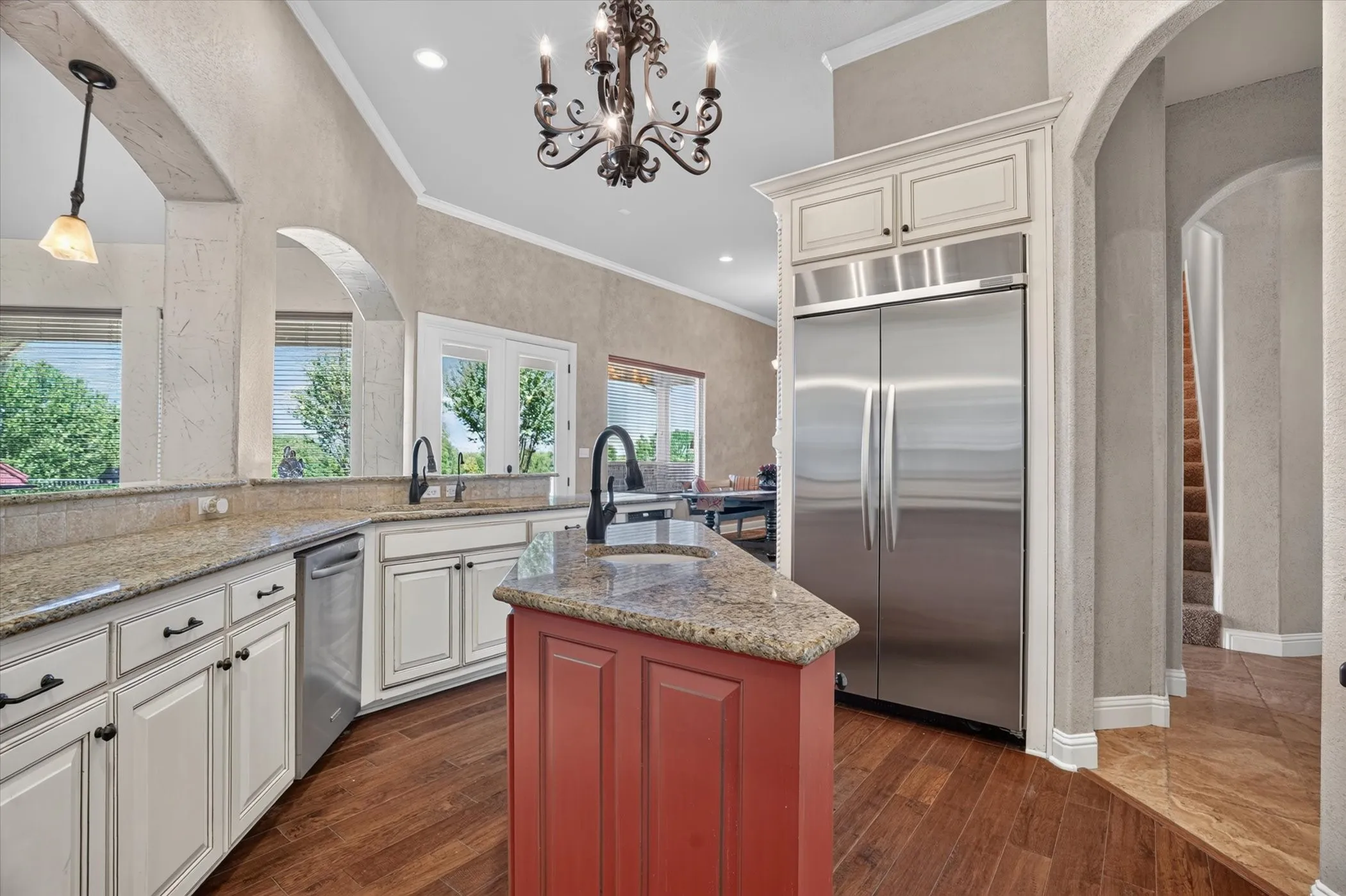 Elegant kitchen featuring cream-painted cabinetry with dark hardware, granite countertops, a custom red-painted island with seating, stainless steel refrigerator and appliances, pendant lighting, arched doorways, recessed ceiling details, and warm wood-finish flooring throughout