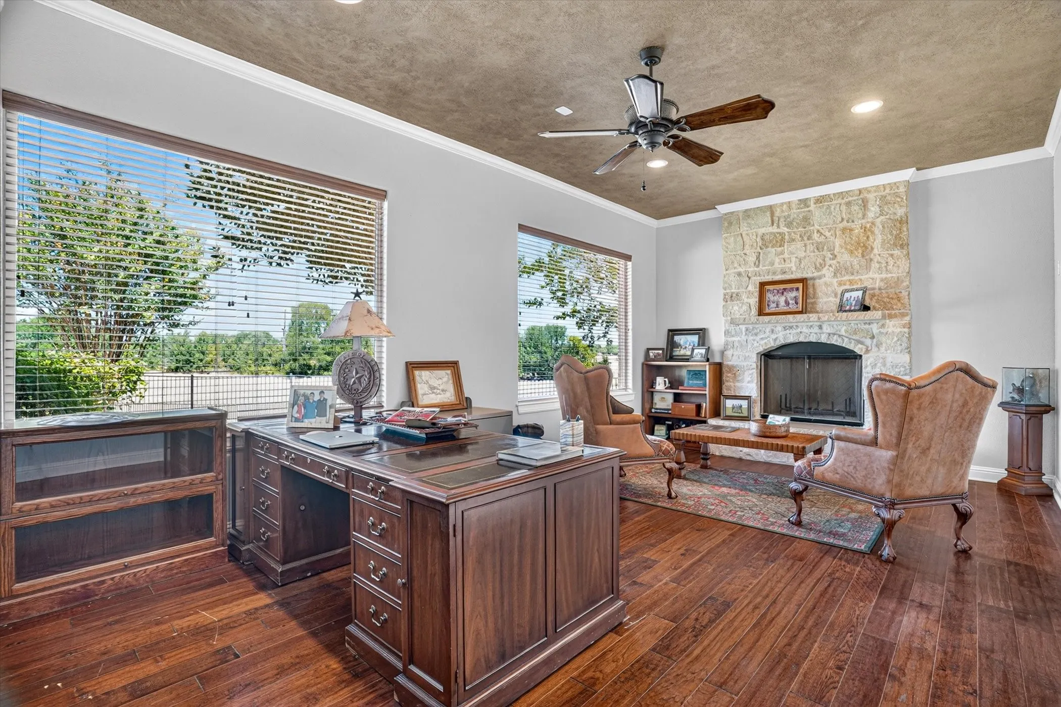 Hardwood flooring, stone-clad fireplace with hearth, ceiling fan, crown molding, and large windows with blinds