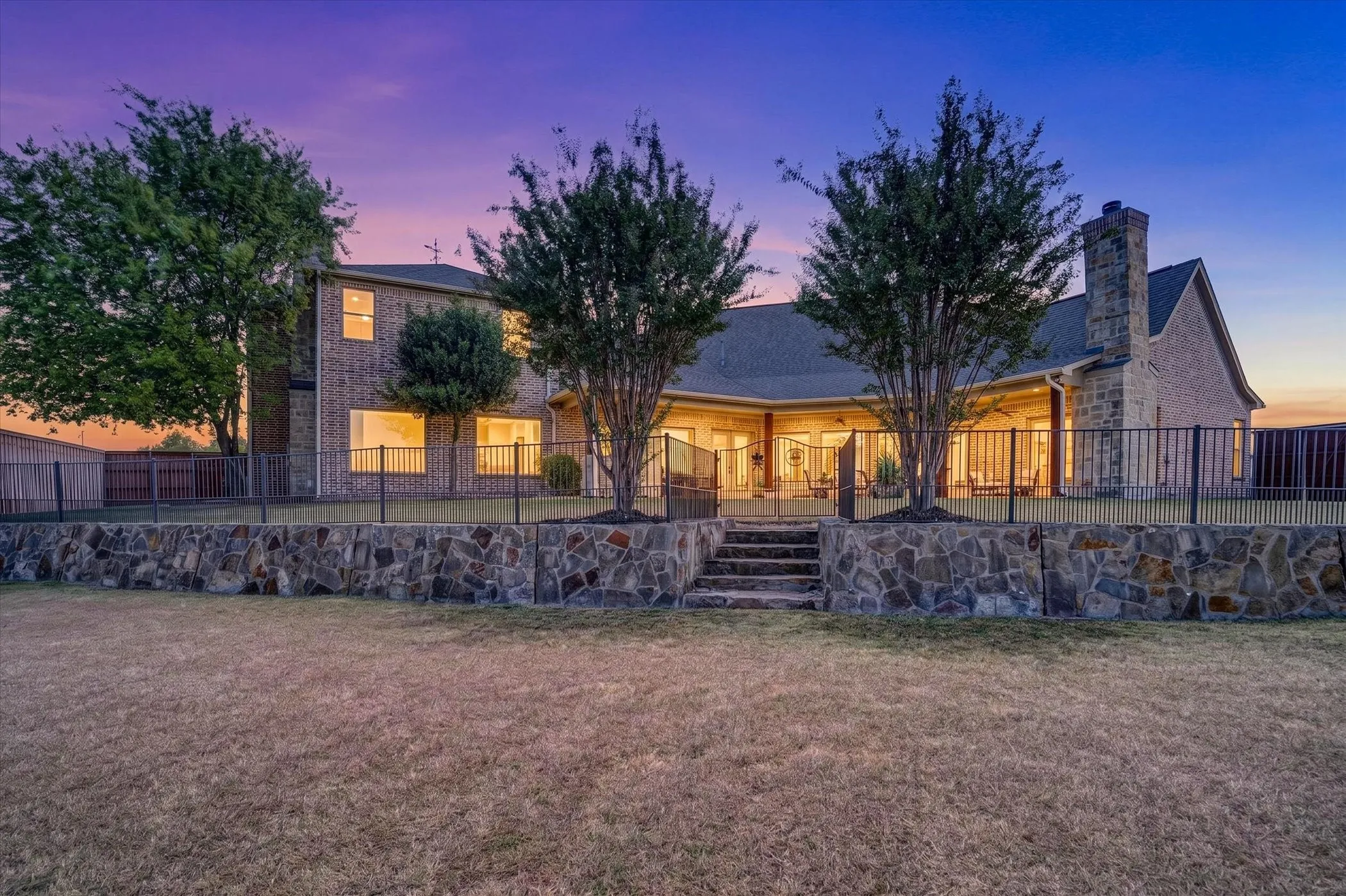 Elegant stone-front residence showcasing arched entryways, multiple illuminated windows, and mature landscaping with specimen trees framing the facade