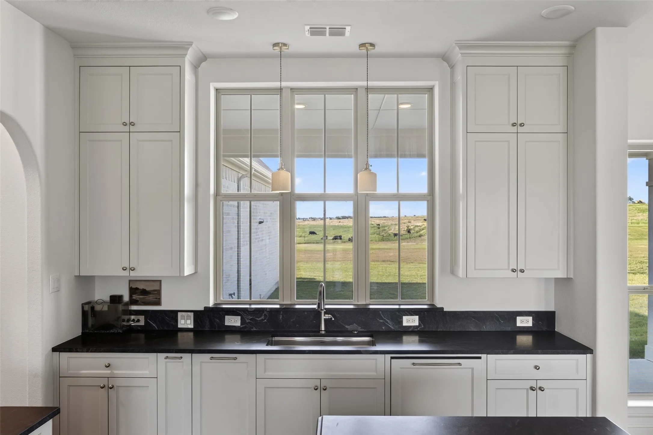 White shaker-style cabinetry with crown molding, dark stone countertops with backsplash, stainless steel sink with a gooseneck faucet, and two pendant lights
