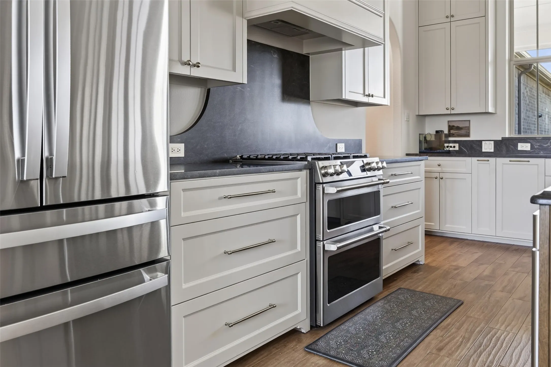Chef's kitchen featuring custom white cabinetry with brushed metal hardware, stainless steel dual-oven range with gas cooktop, stainless steel French-door refrigerator, dark tile backsplash, dark countertops, and wood-finish flooring throughout