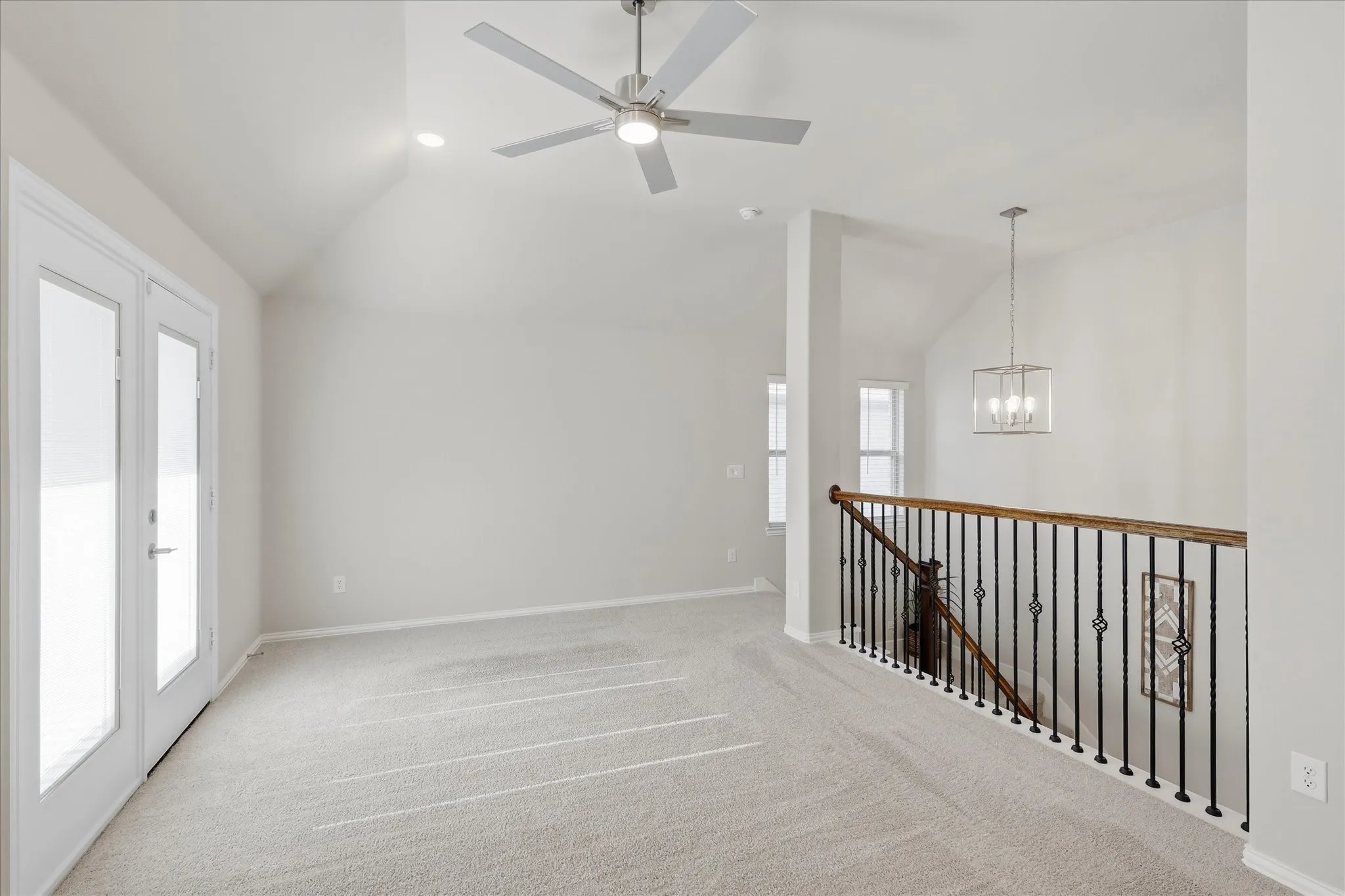 Vaulted ceiling, ceiling fan, recessed lighting, double French doors leading to balcony, and wrought iron railing with wood-finish overlooking staircase