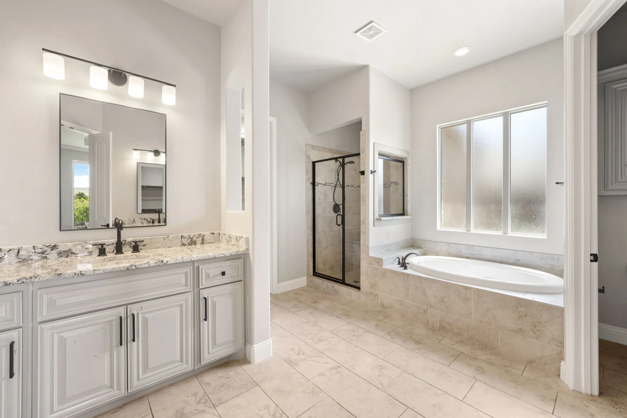 Spacious bathroom featuring a built-in soaking tub, framed glass shower, light wood-look tile flooring, light cabinetry with black hardware, and a vanity with a stone countertop